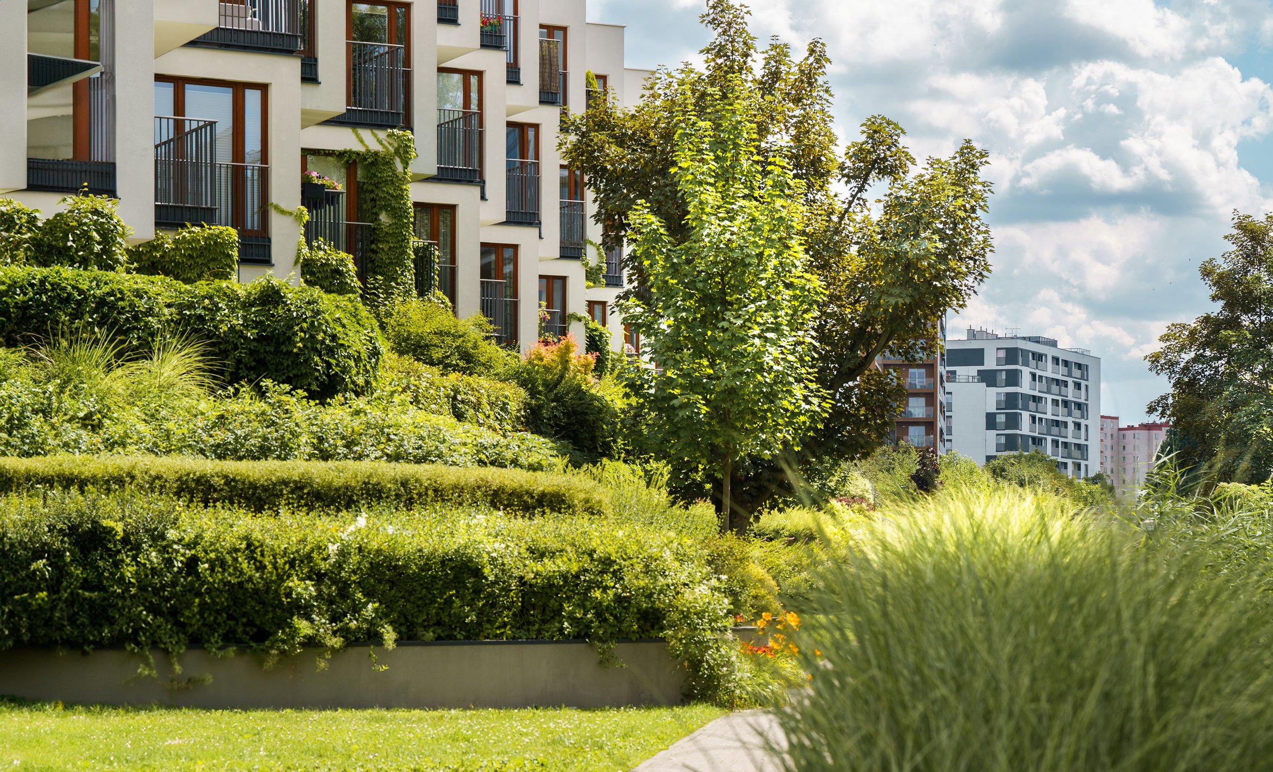 View of a modern apartment building with balconies, surrounded by lush green trees, shrubs, and a manicured lawn on a partly cloudy day.