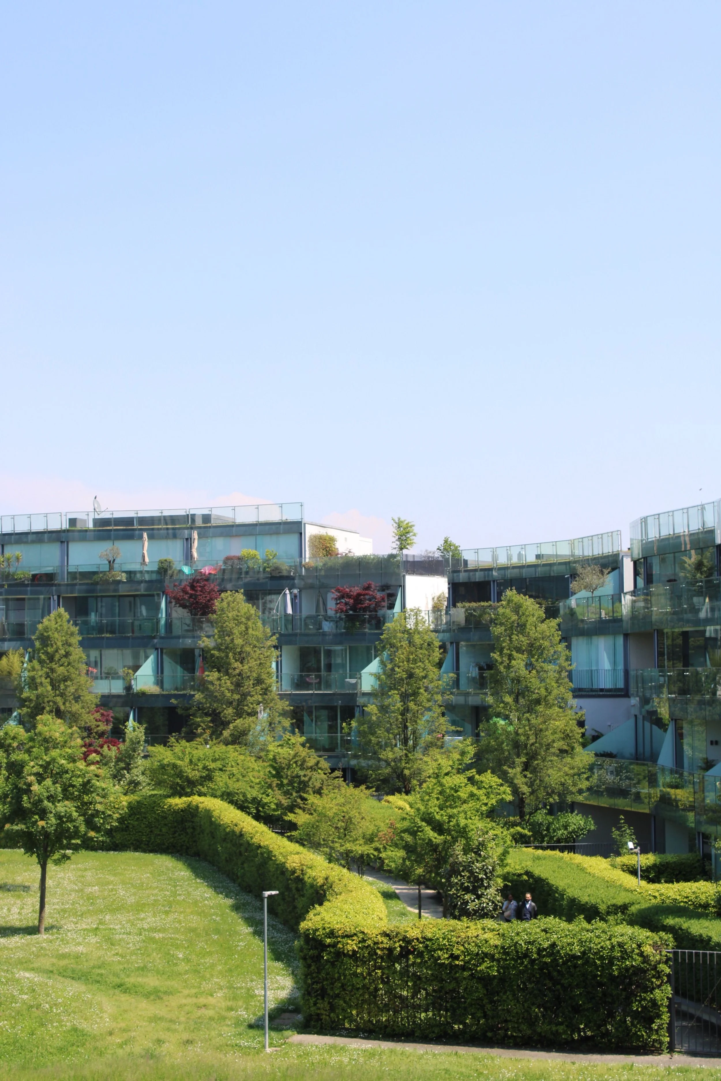 A modern multi-story building with large glass windows, surrounded by lush green trees and a well-maintained park with walking paths and street lights.