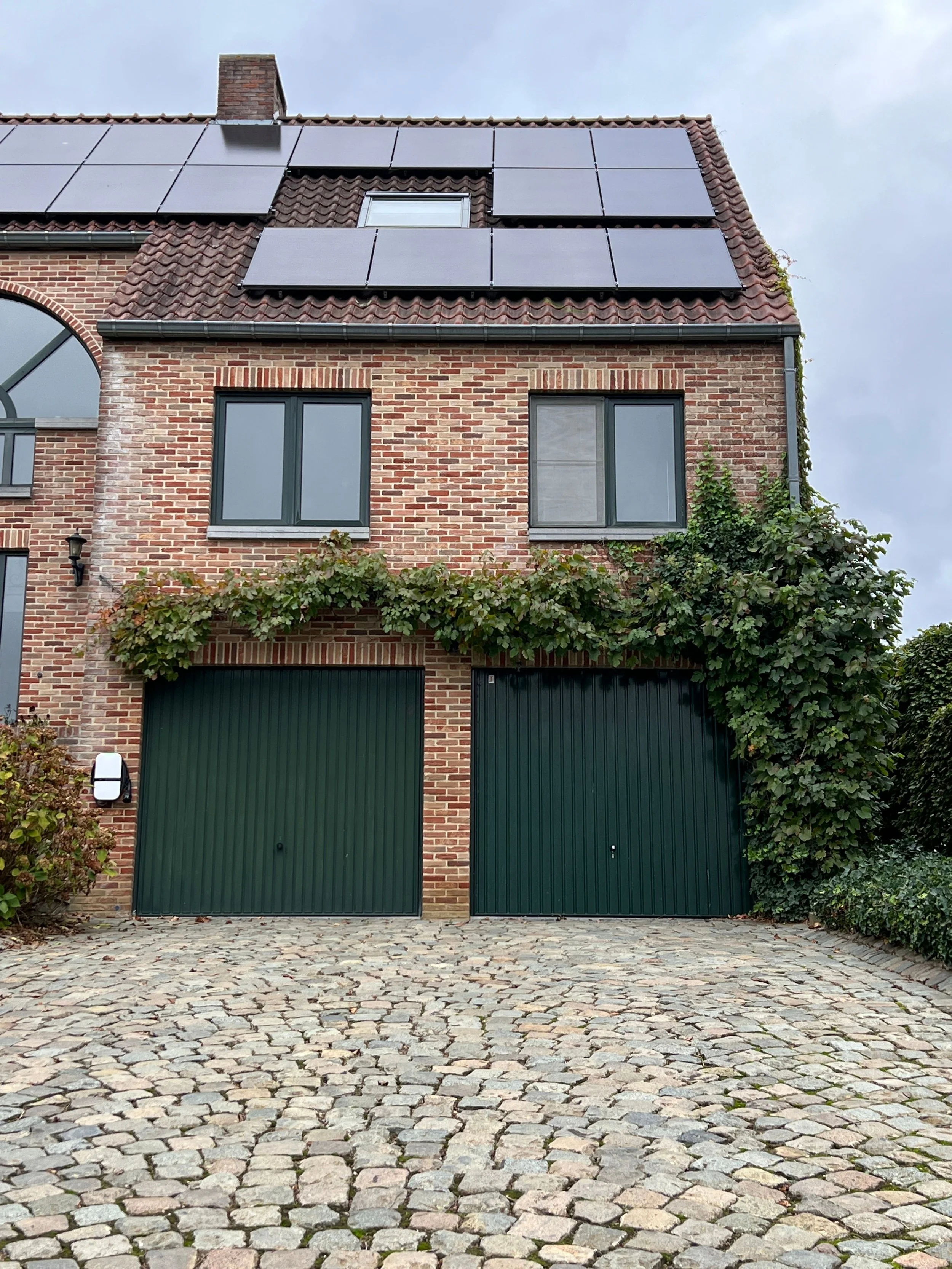 Brick house with two large green garage doors, two windows, and solar panels on the roof, with cobblestone driveway and some greenery.