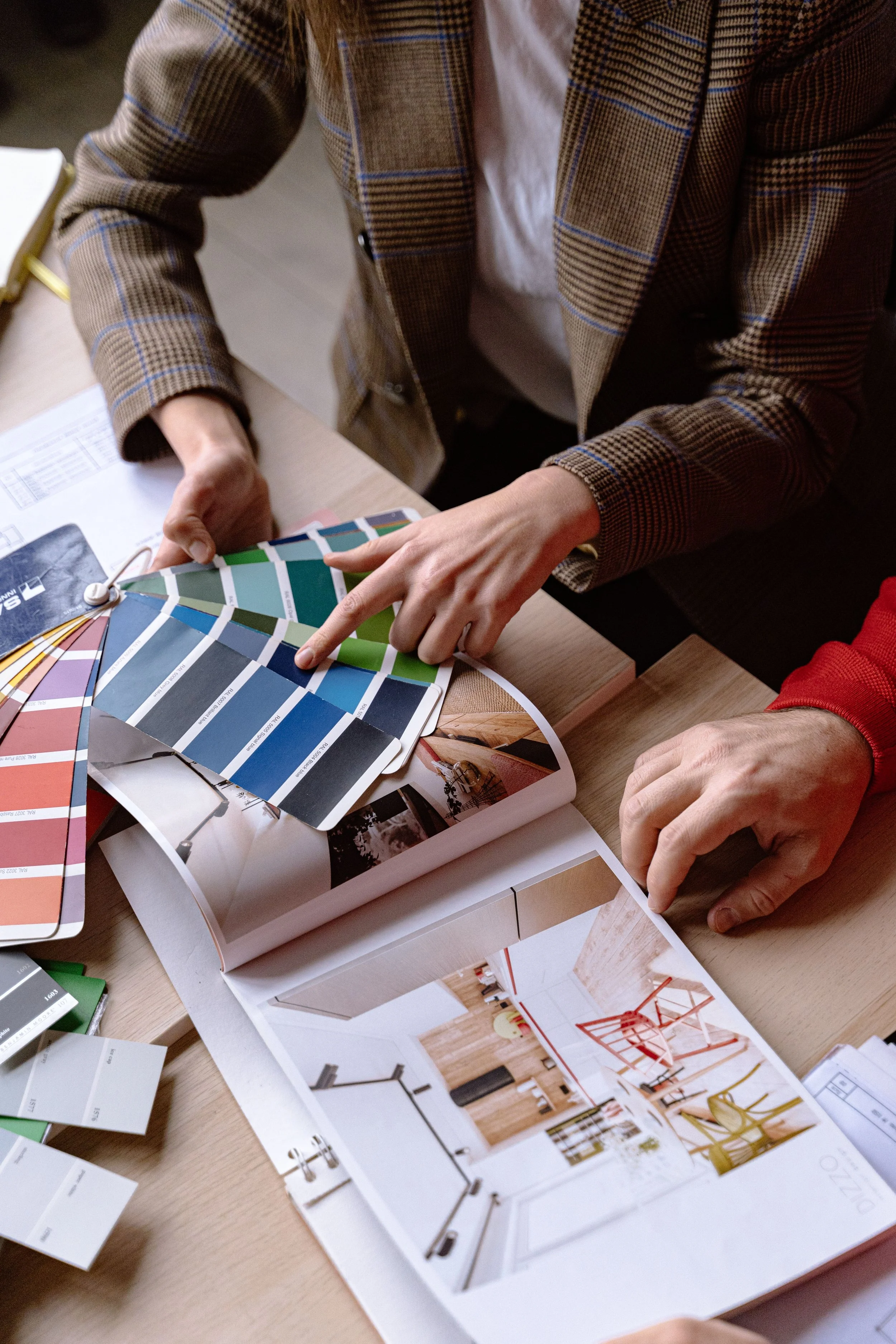 People looking at interior design samples and mood boards on a wooden table, including paint color swatches, magazine pages with furniture and decor ideas.
