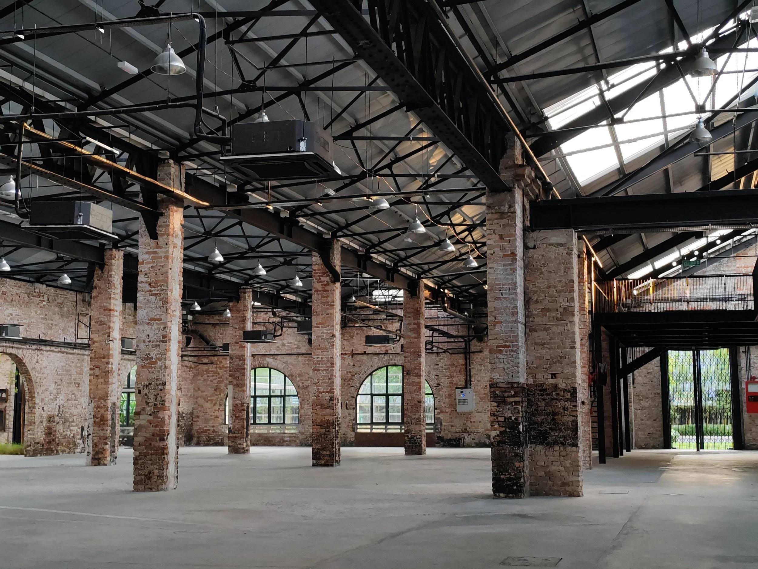 Interior of a large industrial or commercial building with exposed brick pillars, large windows, and metal roofing. The space is empty with a concrete floor and industrial lighting.