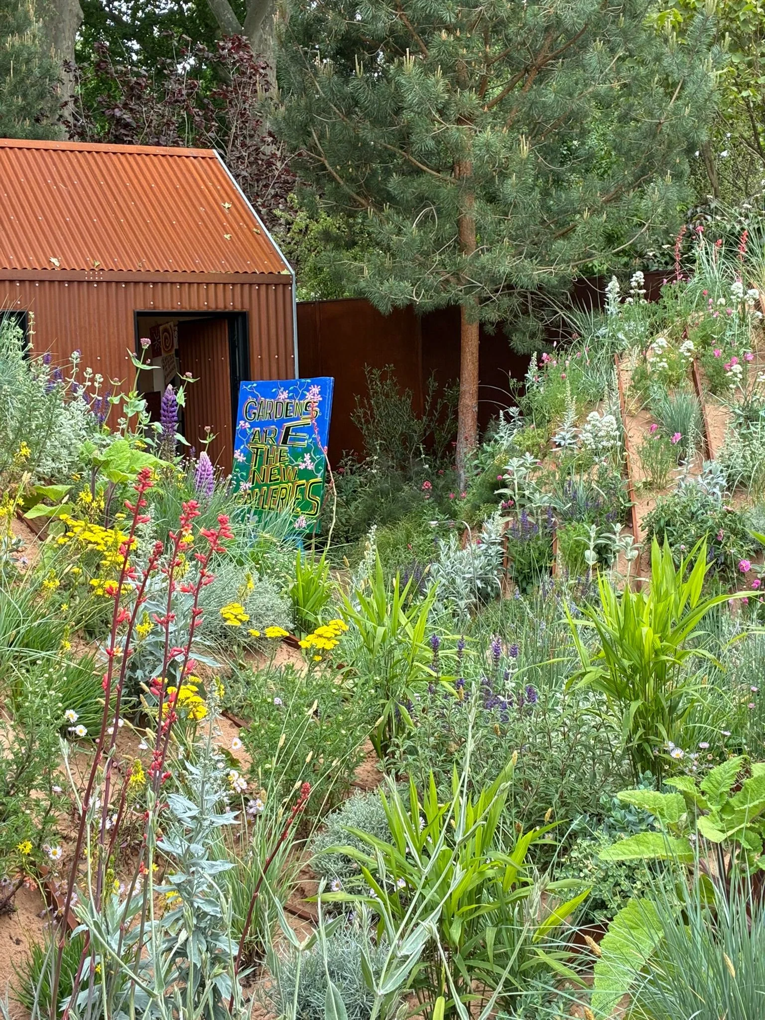 A lush, vibrant garden filled with various colorful flowers and green plants, featuring a small rusty metal shed with an open door and a sign that reads "Gardens are the new galleries." Tall trees and a brown fence surround the garden area.