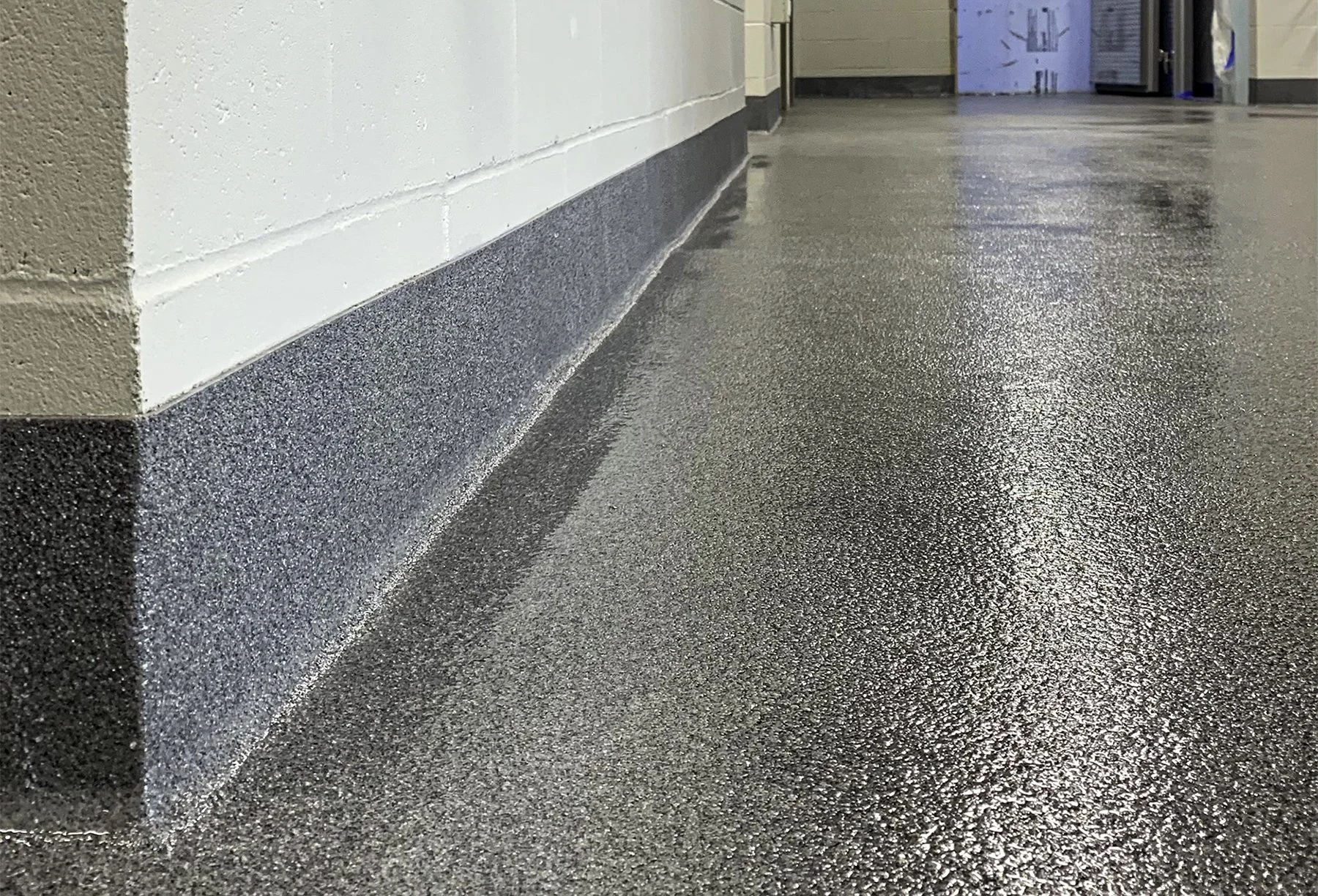 Close-up of a shiny, speckled epoxy floor at the corner of a wall in a commercial or institutional building, with a doorway in the background.
