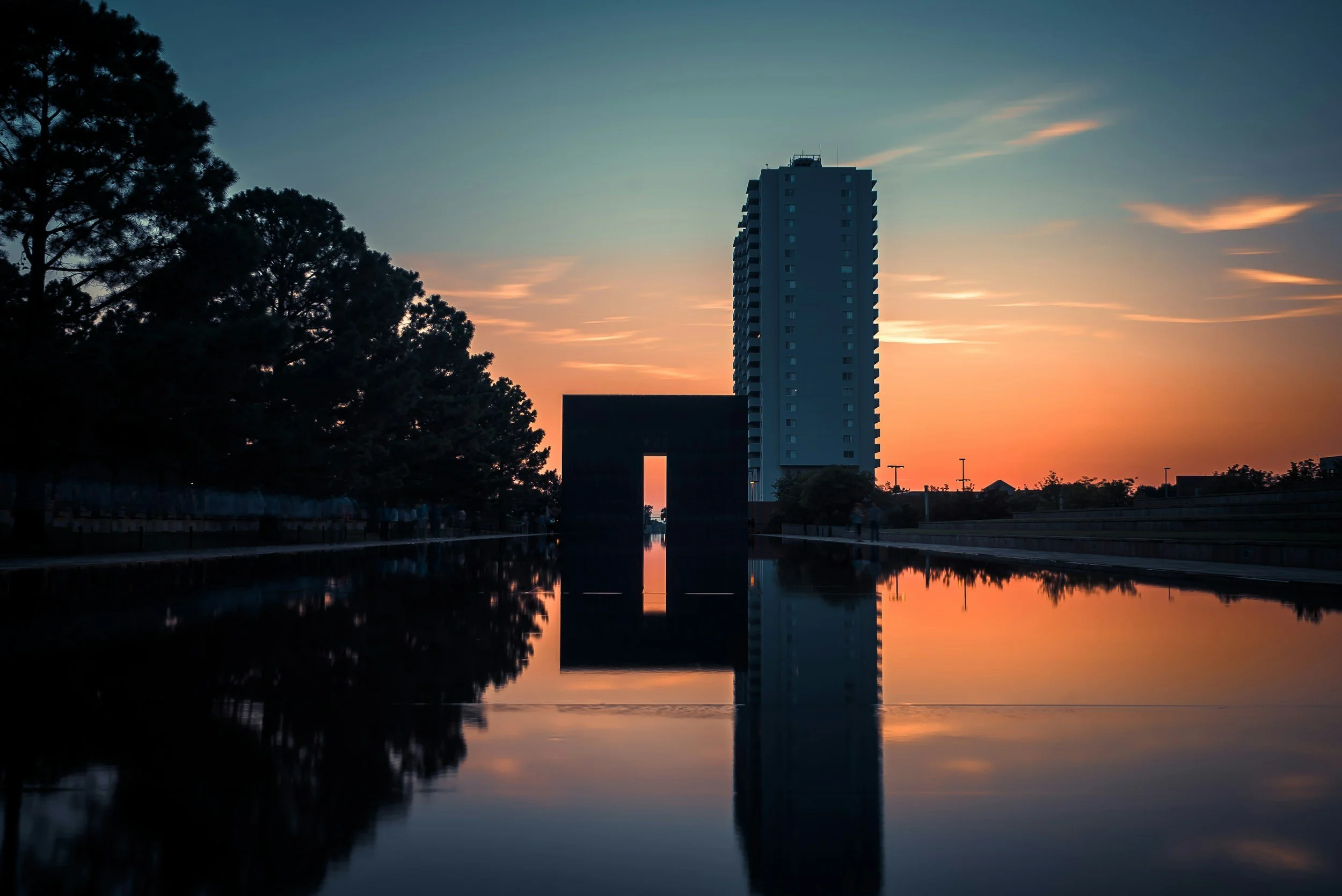 A tall building and trees reflected in a water feature during sunset.