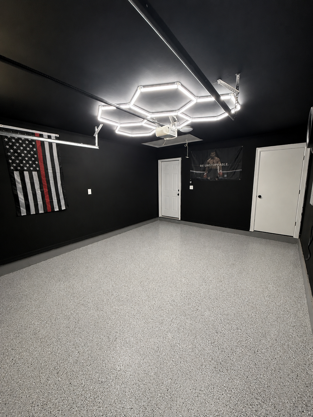 Empty garage with black walls, gray speckled flooring, ceiling-mounted LED light fixture, American flag with a thin red line, and a black banner with a person and the words "Be Unstoppable".