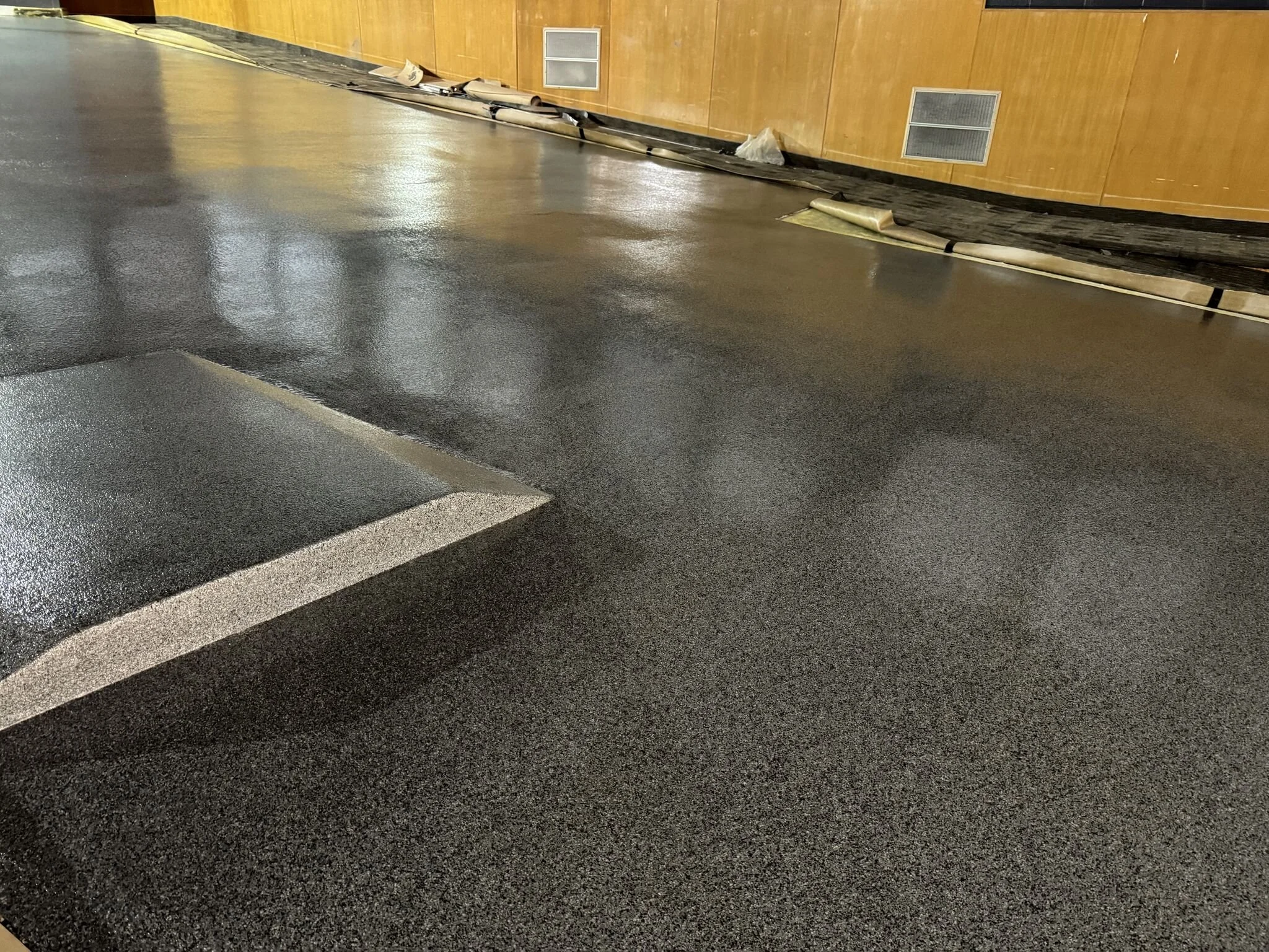 Indoor parking lot with glossy black asphalt floor, yellow wall with vents, and scattered equipment or debris along the base of the wall.