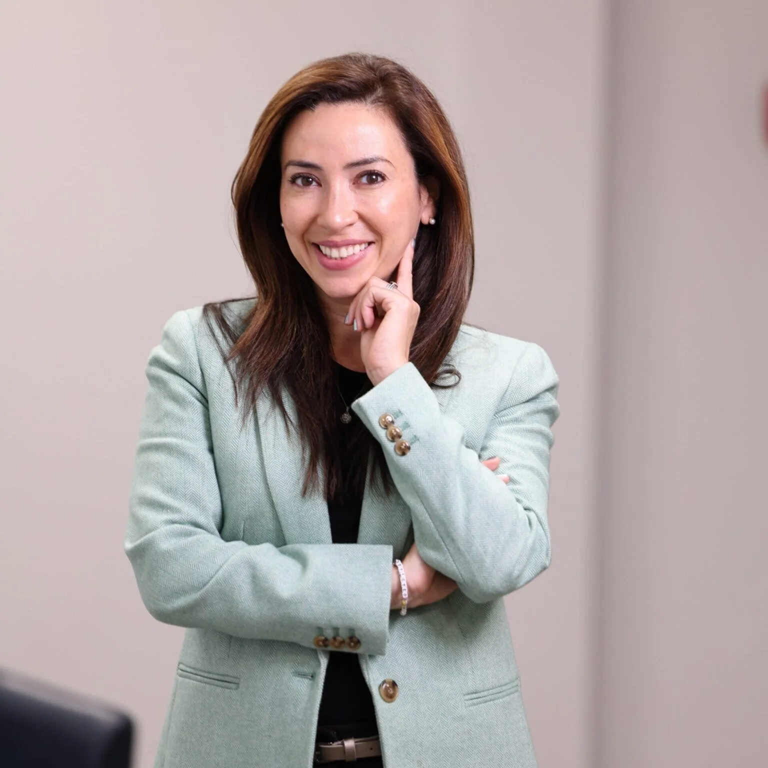 A woman with brown hair in a light green blazer smiling and touching her face with her right hand, standing against a plain wall.