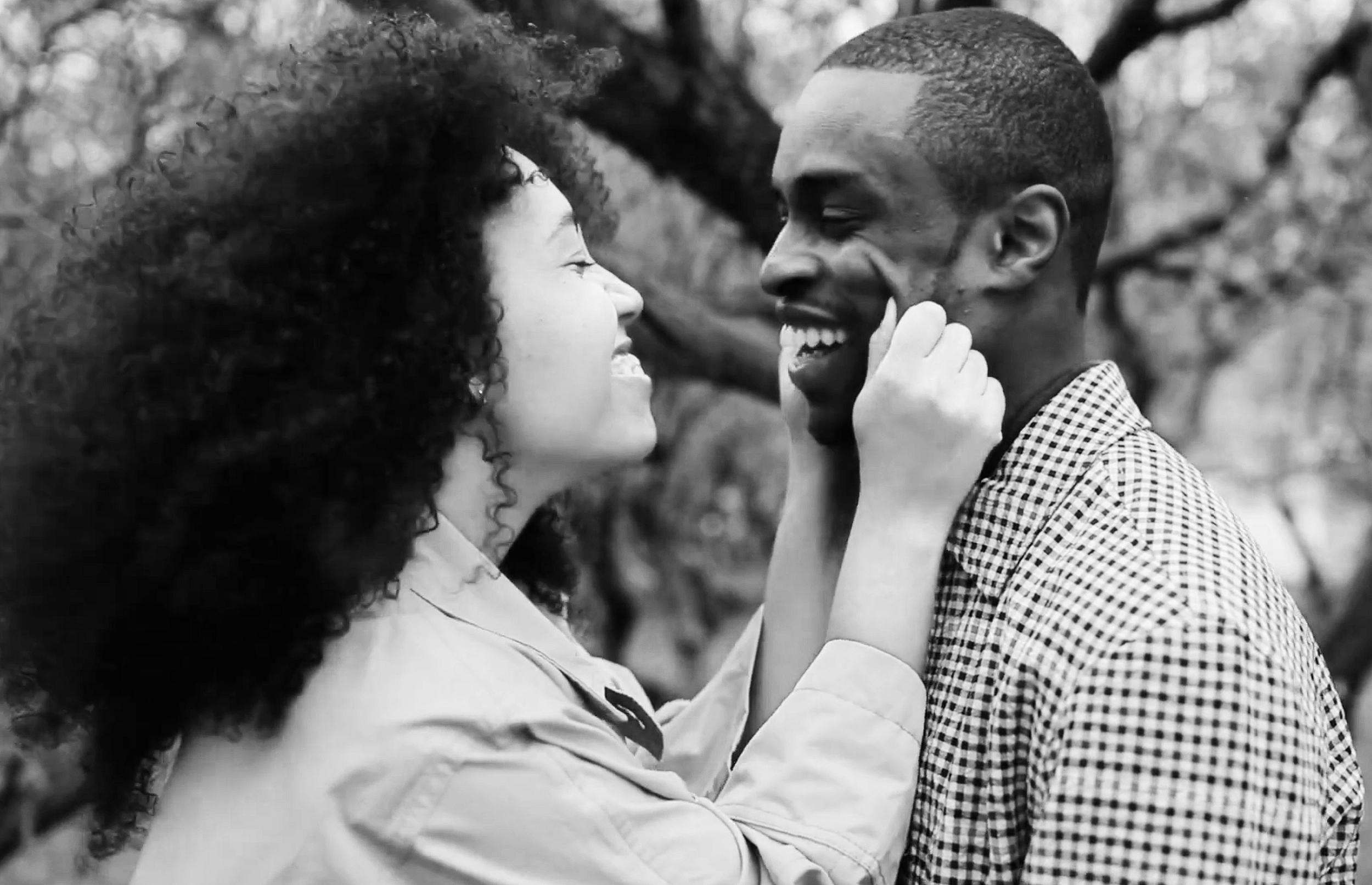 A black-and-white photo of a smiling Black couple looking into each other's eyes, close-up, outdoors with blurred tree branches in the background.