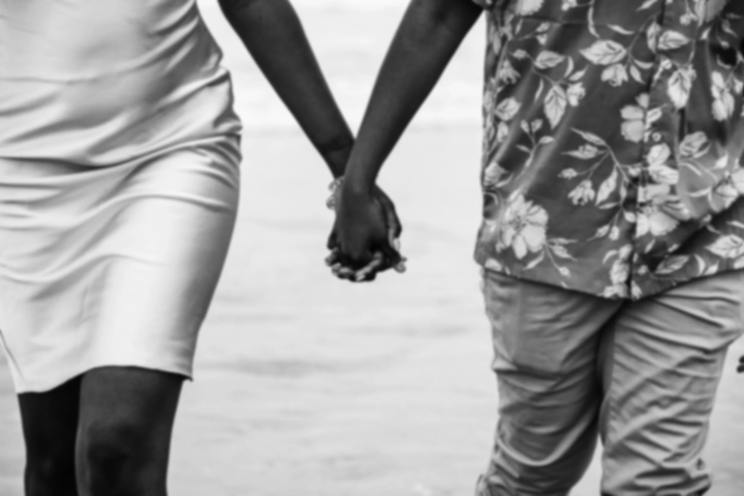 Close-up of a couple holding hands, wearing casual shorts and patterned shirt, walking together on a beach in black and white.