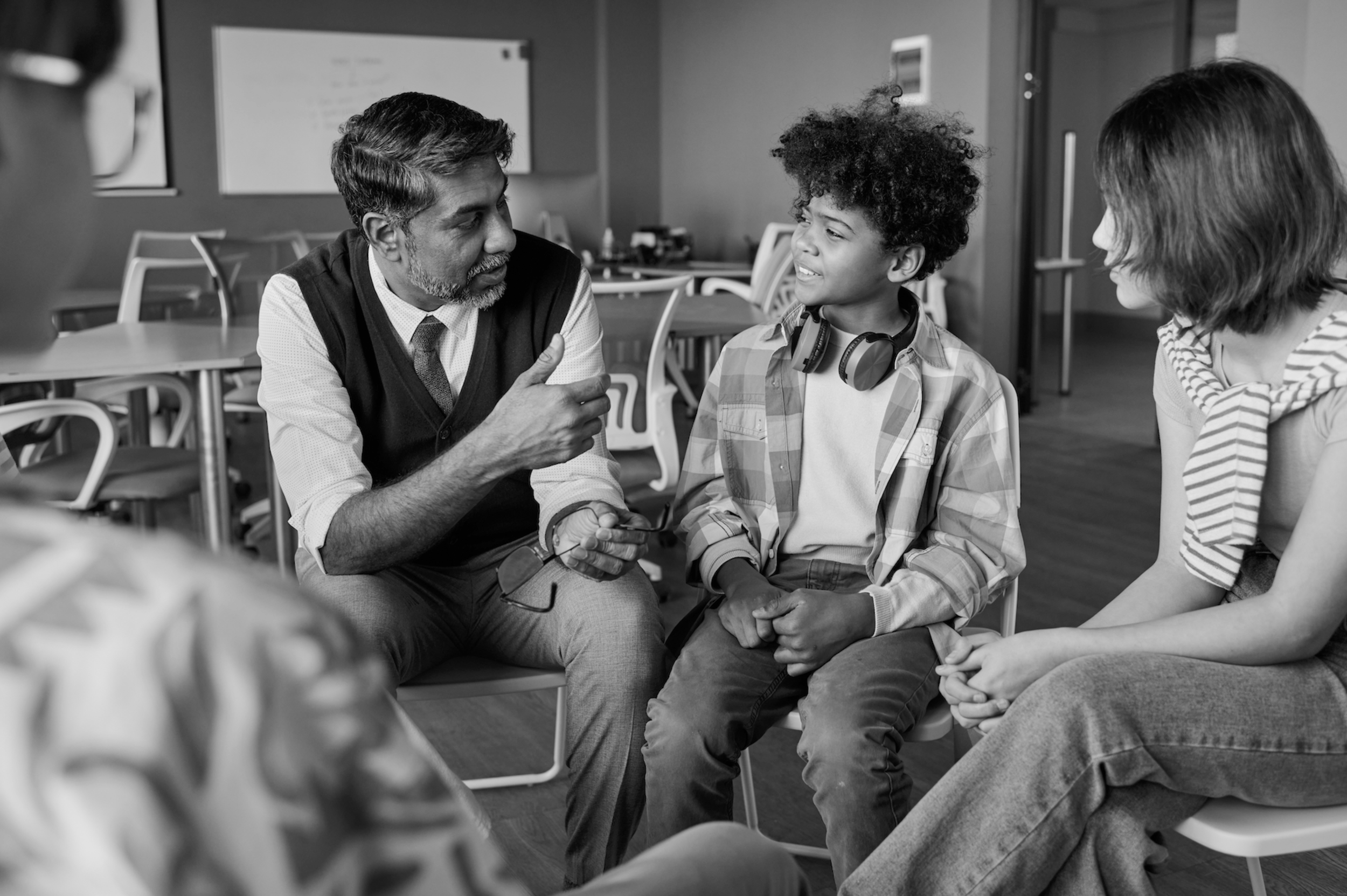 A man with gray beard and a woman with short hair are sitting on chairs, holding hands, and speaking to a young boy with curly hair who is smiling. The scene takes place in a room with tables and chairs in the background.