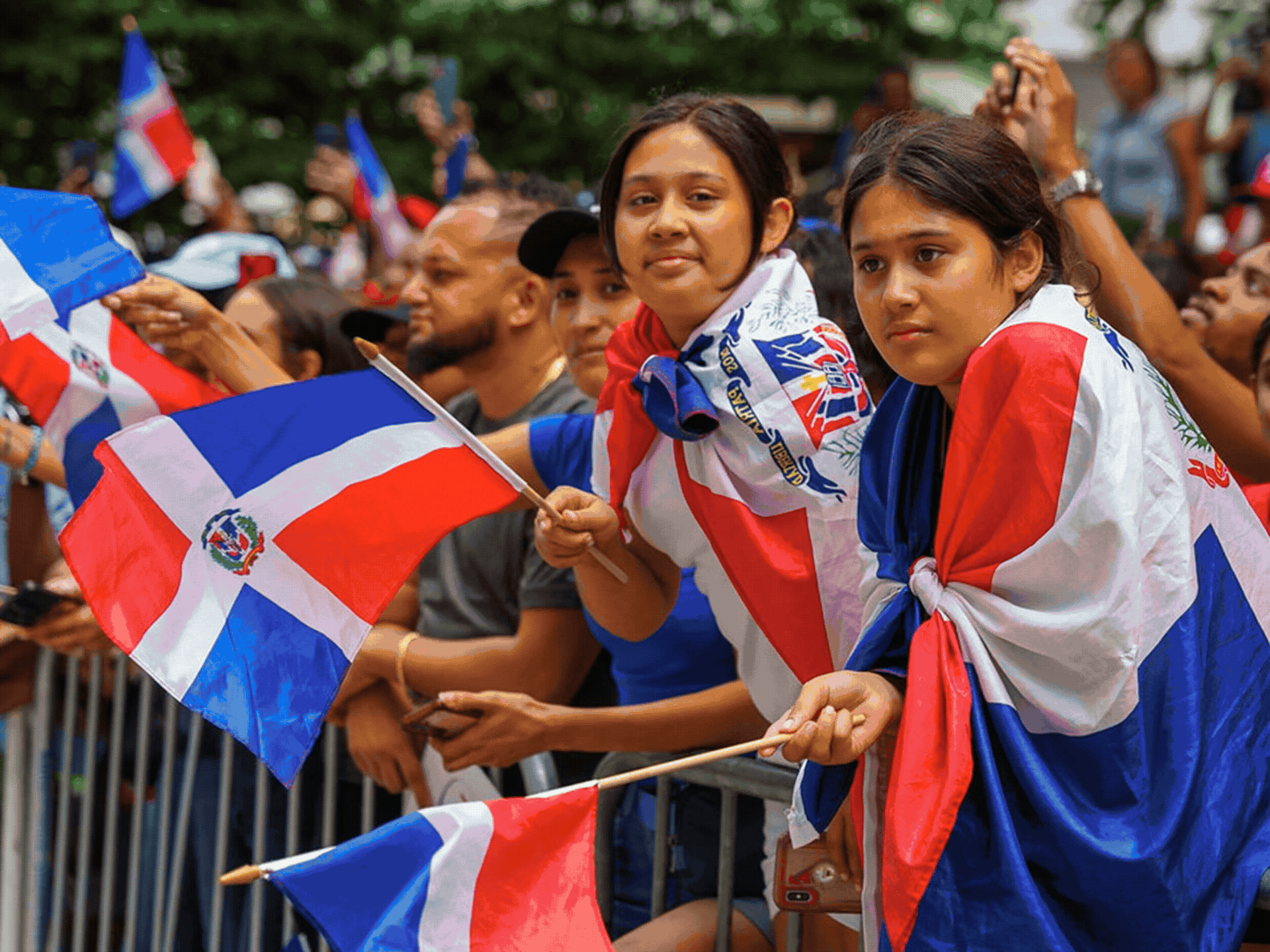 People in a crowd holding and waving flags with red, white, and blue colors, and some flags with emblem designs, during an outdoor event.