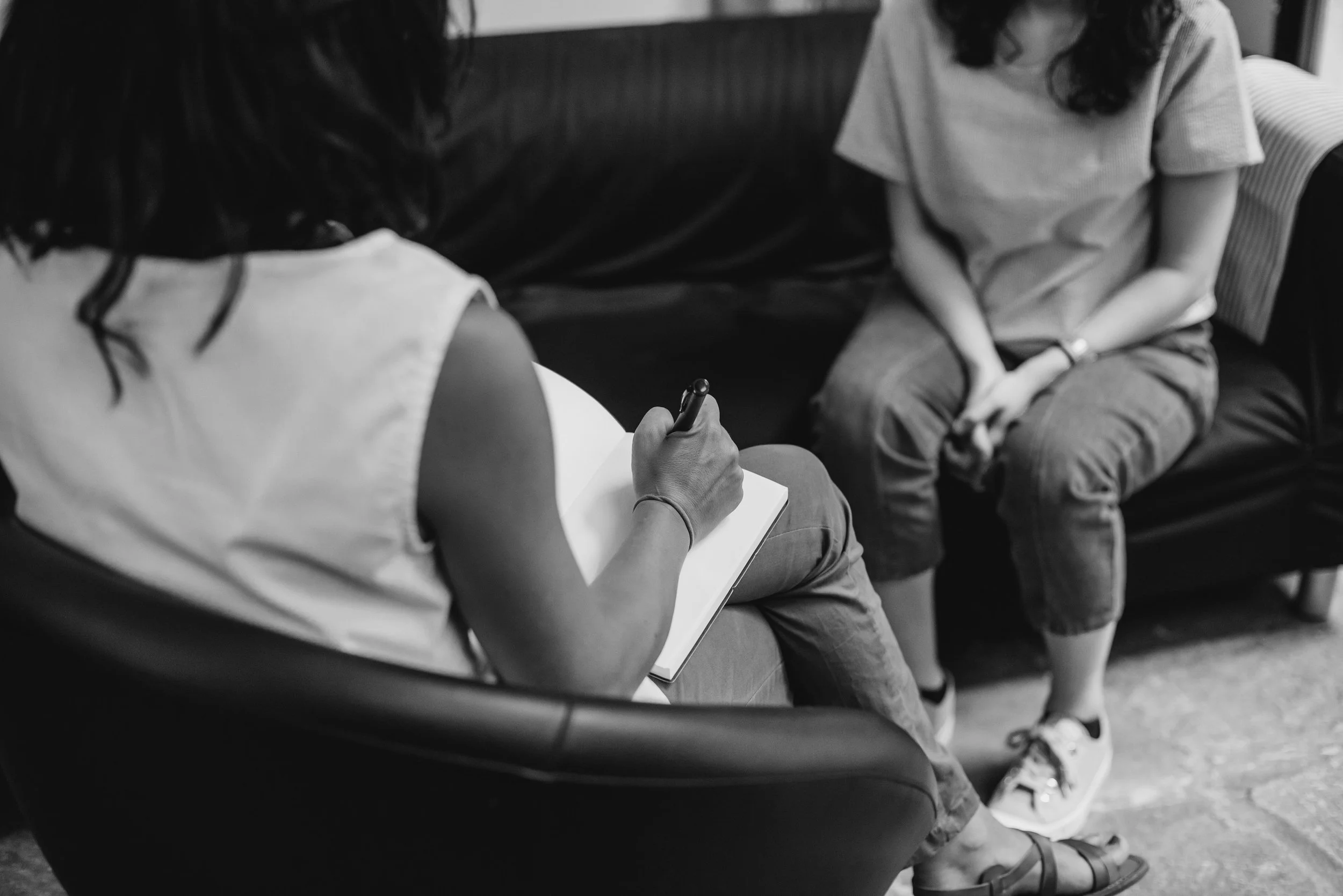 Two people sitting on chairs, one writing on a notepad and the other sitting with hands clasped, both facing each other. The image is in black and white.