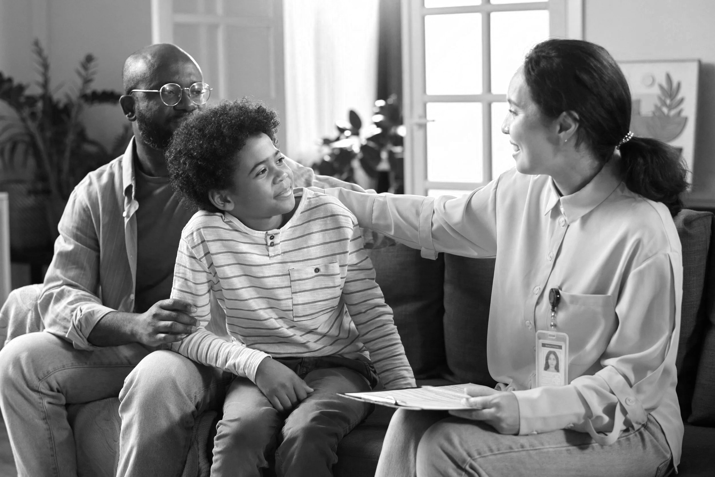 A woman in a medical coat smiling and talking to a young boy in a striped shirt sitting on a man's lap. The man is also smiling, and they are in a room with large windows and plants.