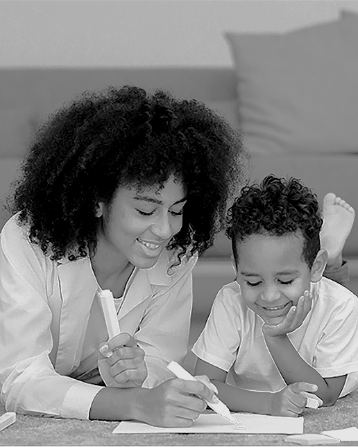 A woman and a young boy sitting on the floor, smiling as they draw or write on a piece of paper.