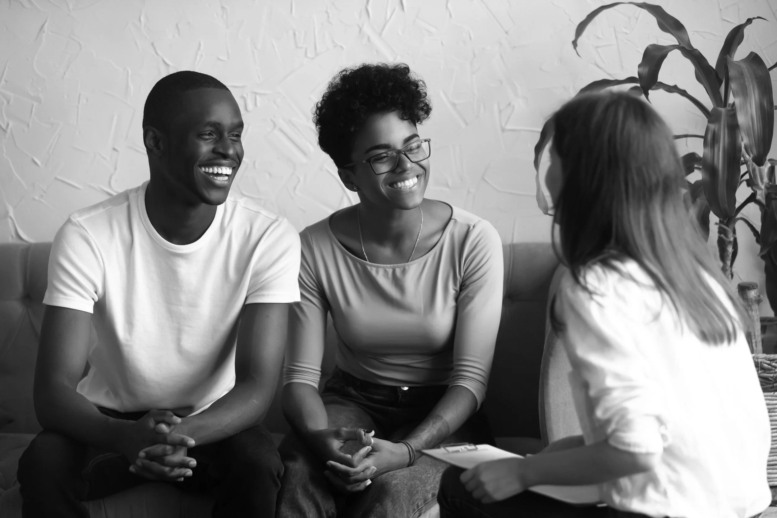 Four people are sitting together indoors, smiling and engaging in conversation. Three of them are facing the camera: a smiling young man with short hair, a young woman with glasses and curly hair, and another person's head visible. The fourth person,