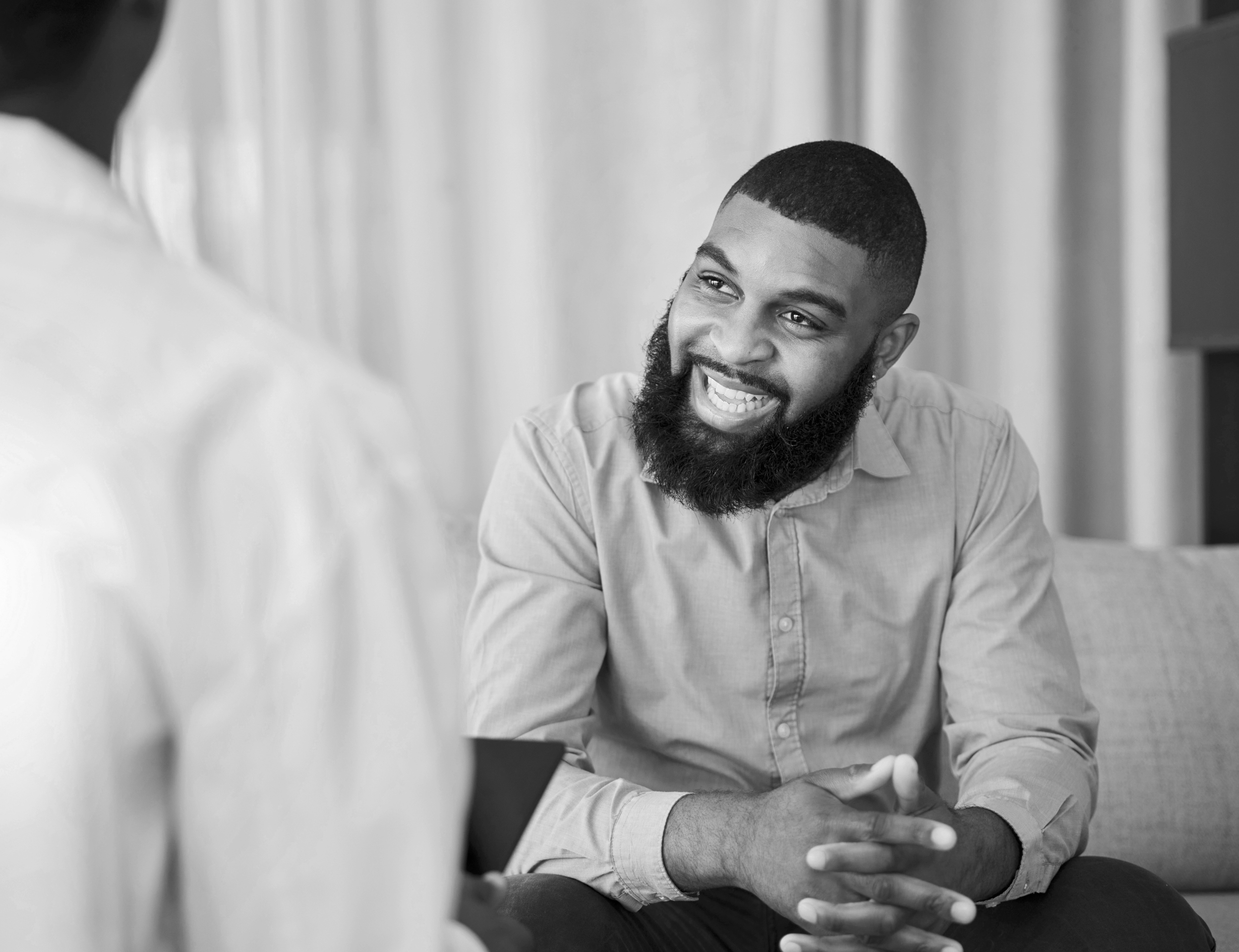 A smiling man with a beard and short hair, wearing a light-colored shirt, is engaged in conversation with another person, partially visible and out of focus, in an indoor setting with a neutral background.