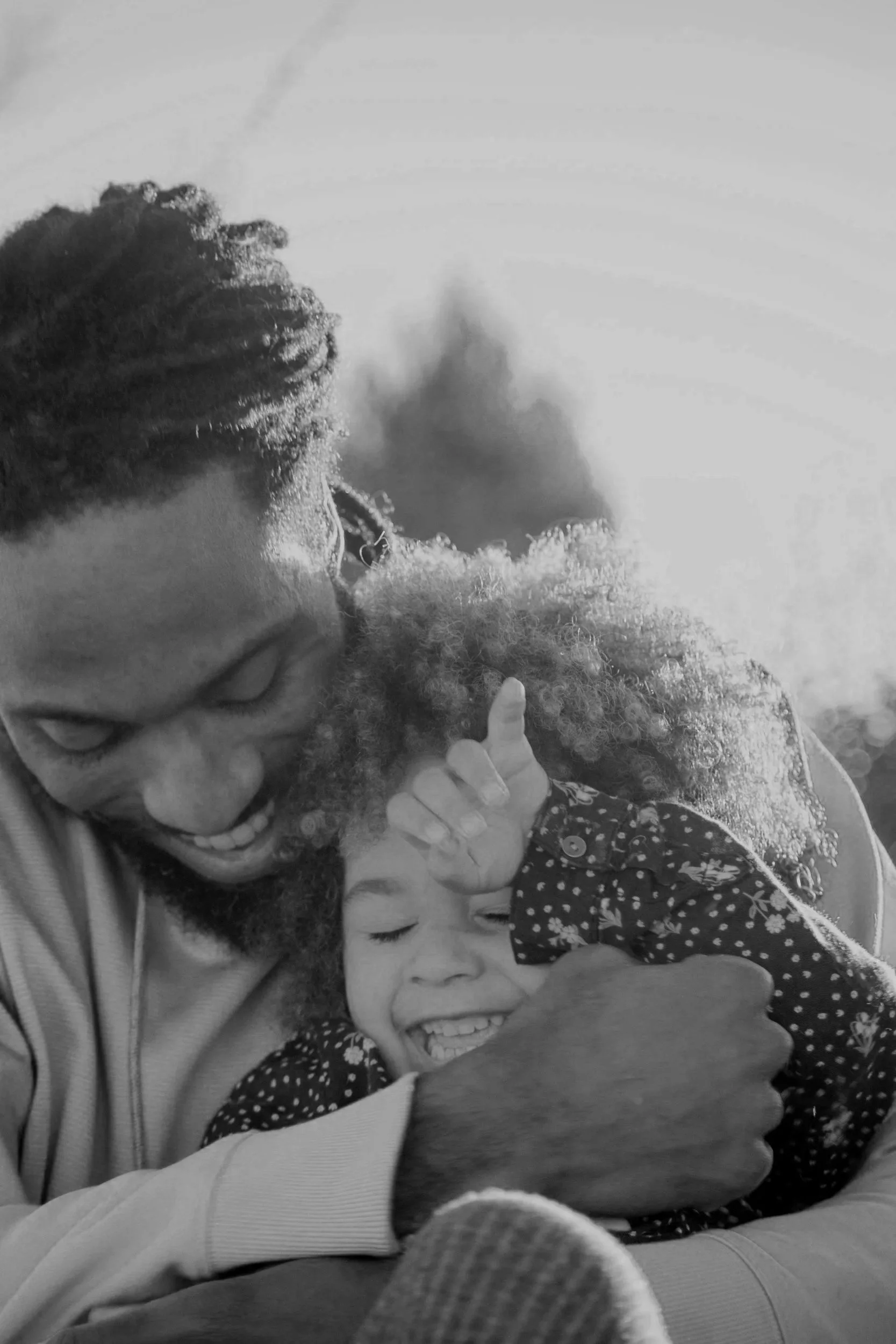 A man with glasses and a beard, smiling, is hugging two children, a boy and a girl, in a happy family moment. The boy is smiling widely, and the girl has her eyes closed, wearing a polka dot dress.