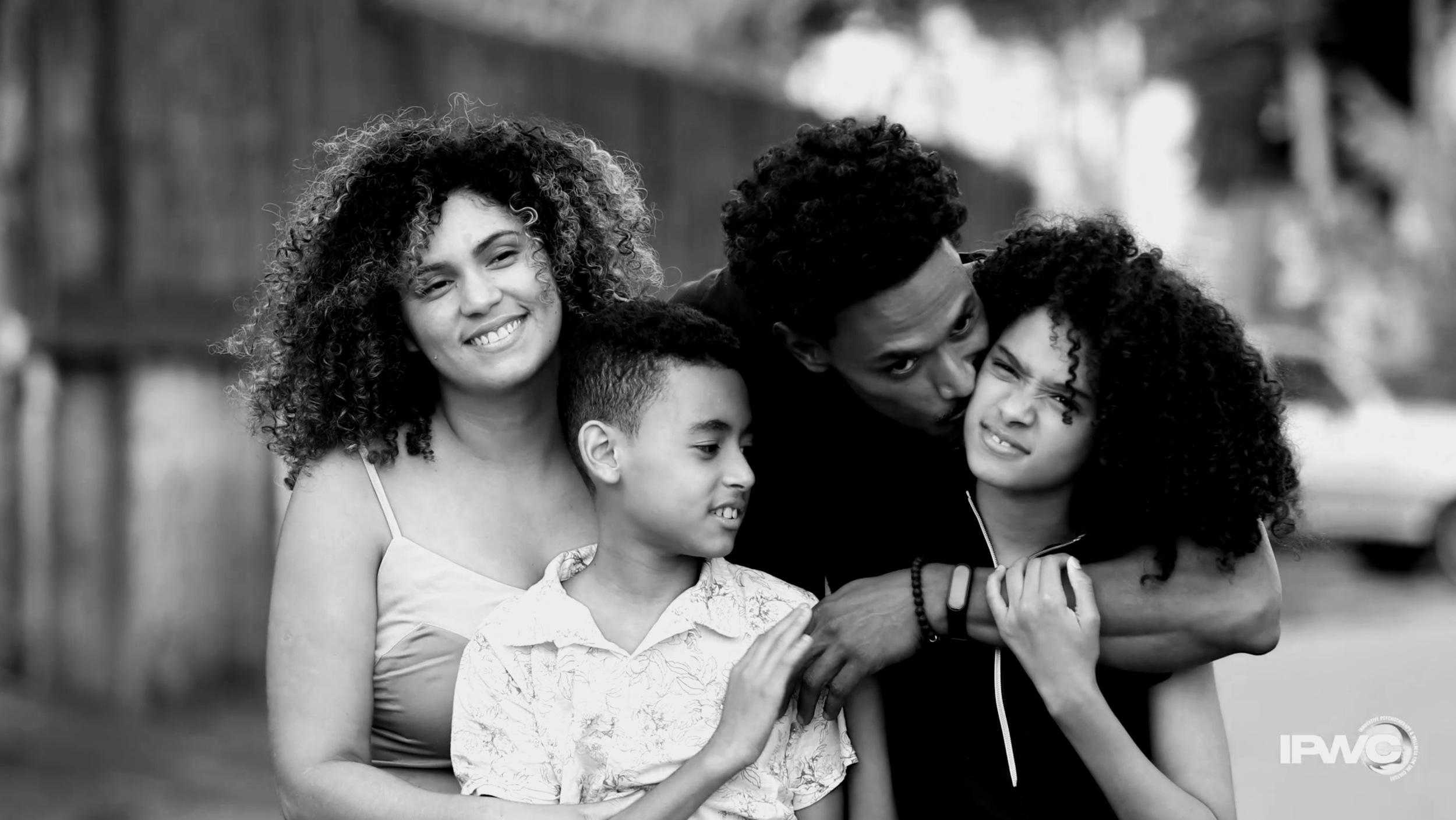 A happy mixed-race family of five outdoors. The mother, father, and three children are hugging and smiling at the camera, with a wooden fence and trees in the background.