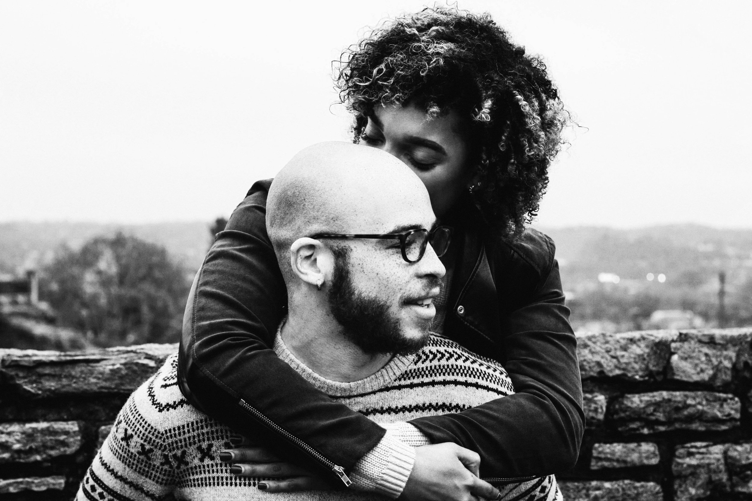 A black-and-white photo of a woman with curly hair hugging a man with a beard, bald head, and glasses. They are outdoors in front of a stone wall.