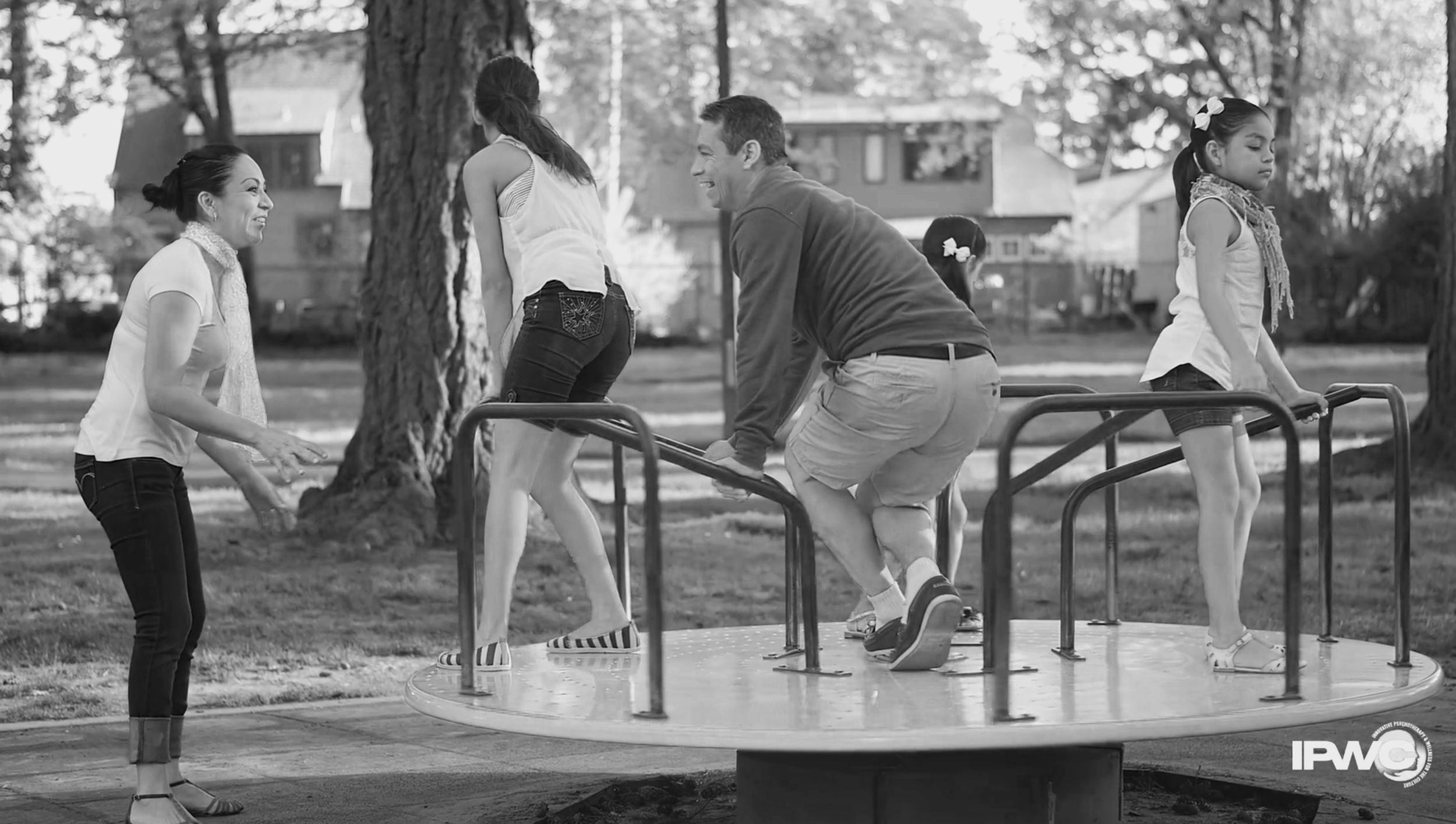 People at a playground, with two young girls on a spinning ride and two adults nearby, in a park with trees and houses in the background.