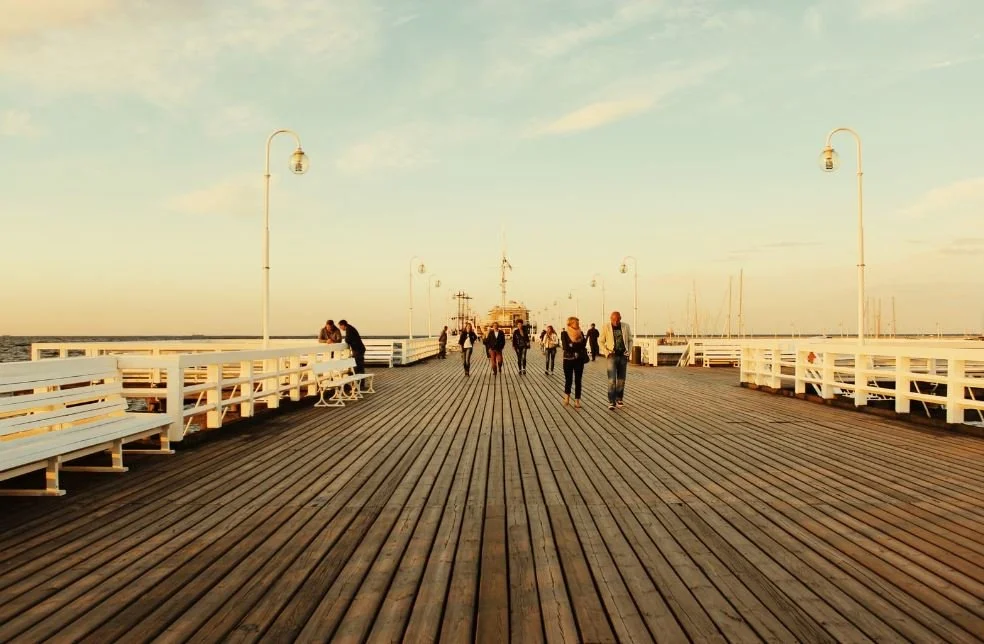 People walking on a wooden pier with benches and lampposts, leading toward the water and a boat in the distance during sunset.