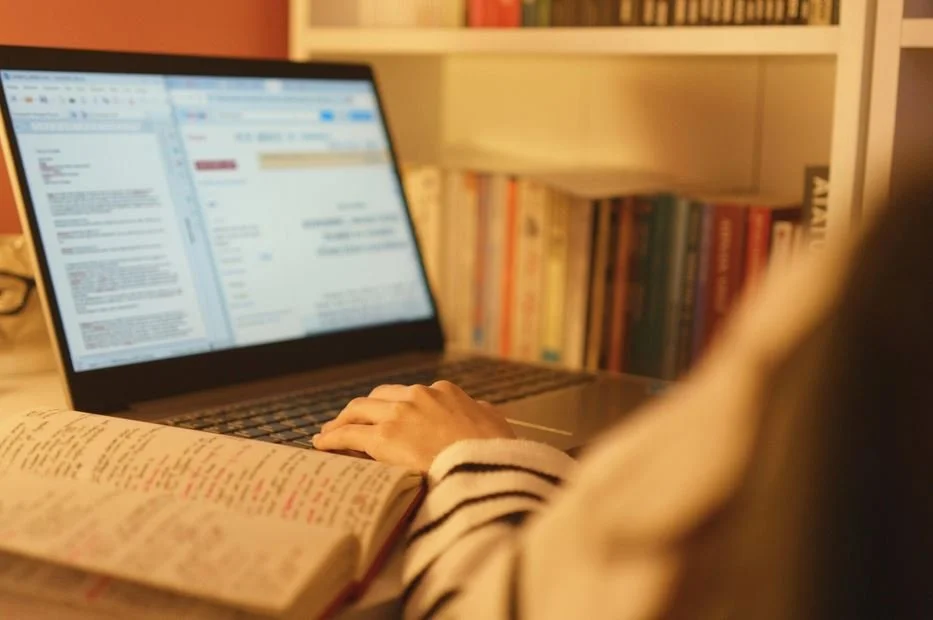 Person studying with an open book and using a laptop at a desk with a bookshelf in the background.