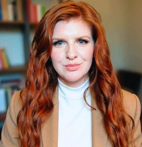 A woman with long wavy red hair, blue eyes, wearing a white turtleneck and tan blazer, in front of a blurred bookshelf background.