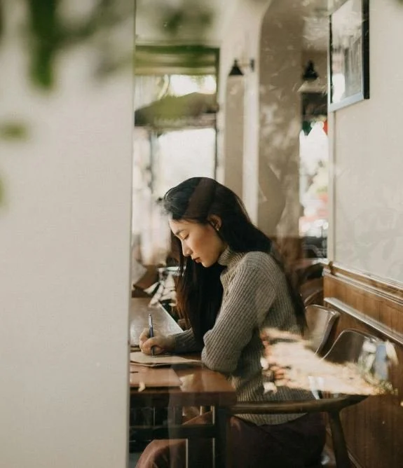Woman sitting at a cafe table, writing in a notebook, with windows and interior decor in the background.
