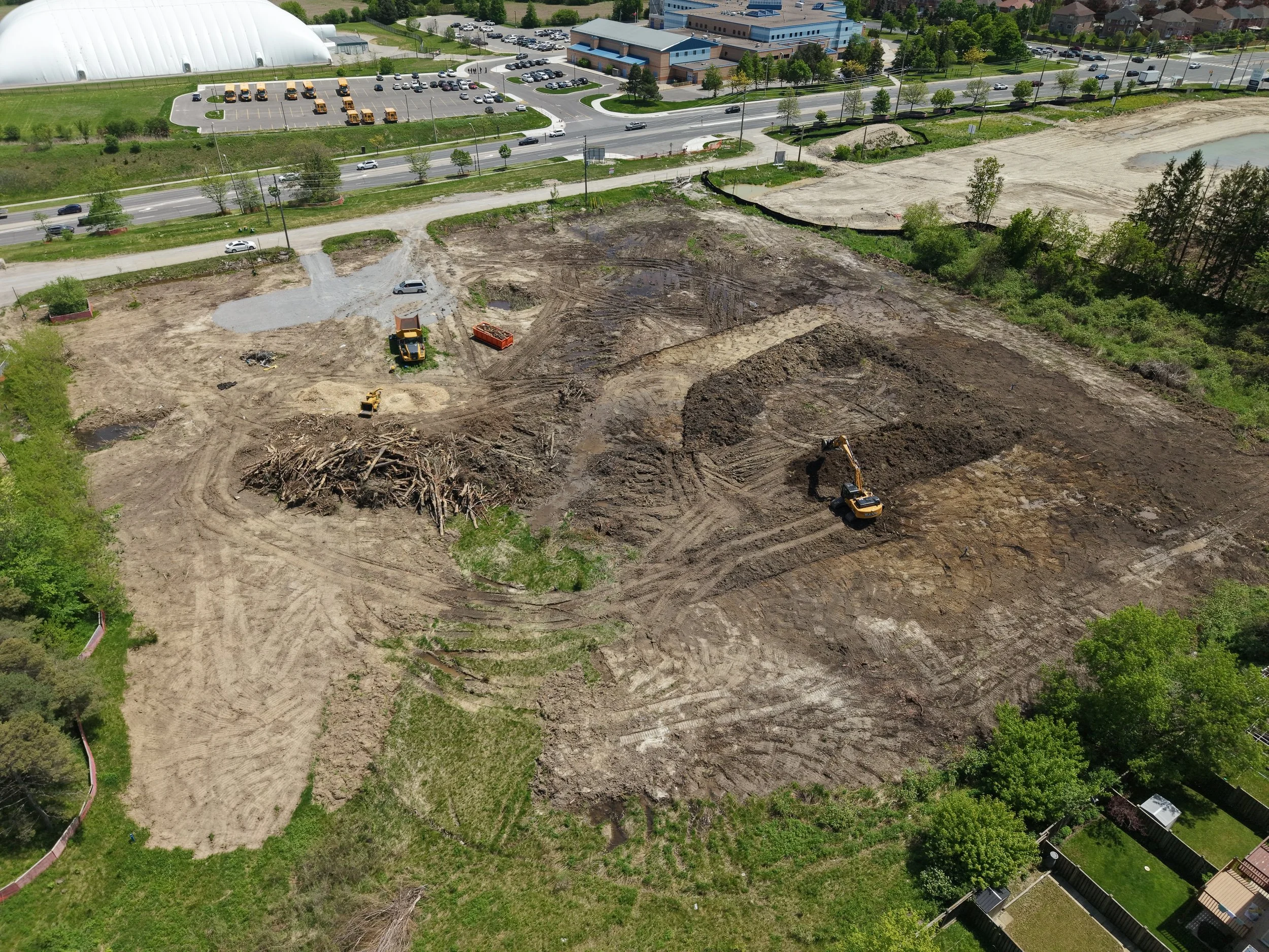 An aerial view of a construction site with cleared land, construction equipment, and some patches of water, adjacent to a road and a parking lot with buses and vehicles, and a building with trees on a sunny day.