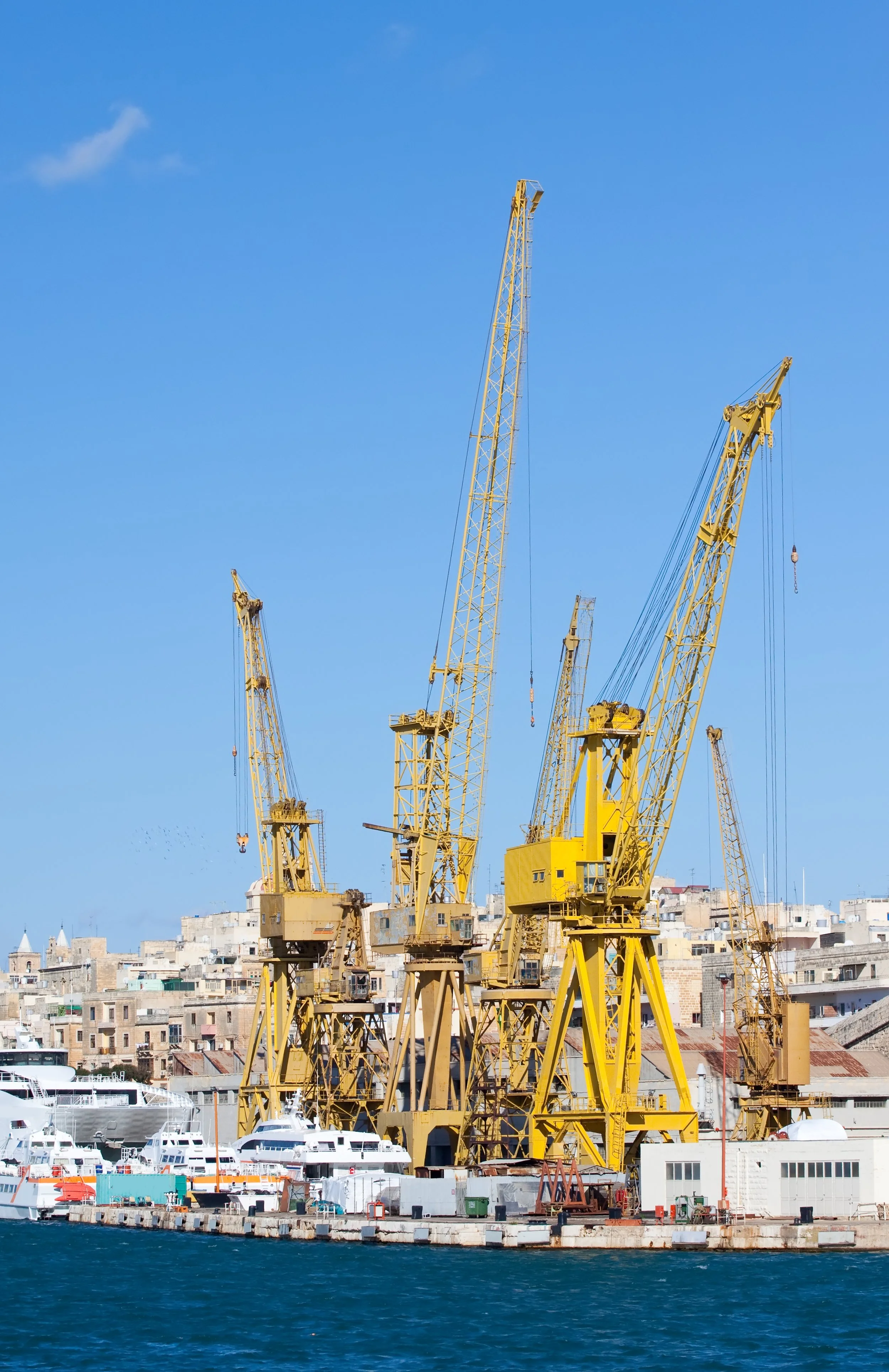 View of a harbor with multiple yellow cranes, boats docked along the water, and a cityscape with buildings in the background under a blue sky.