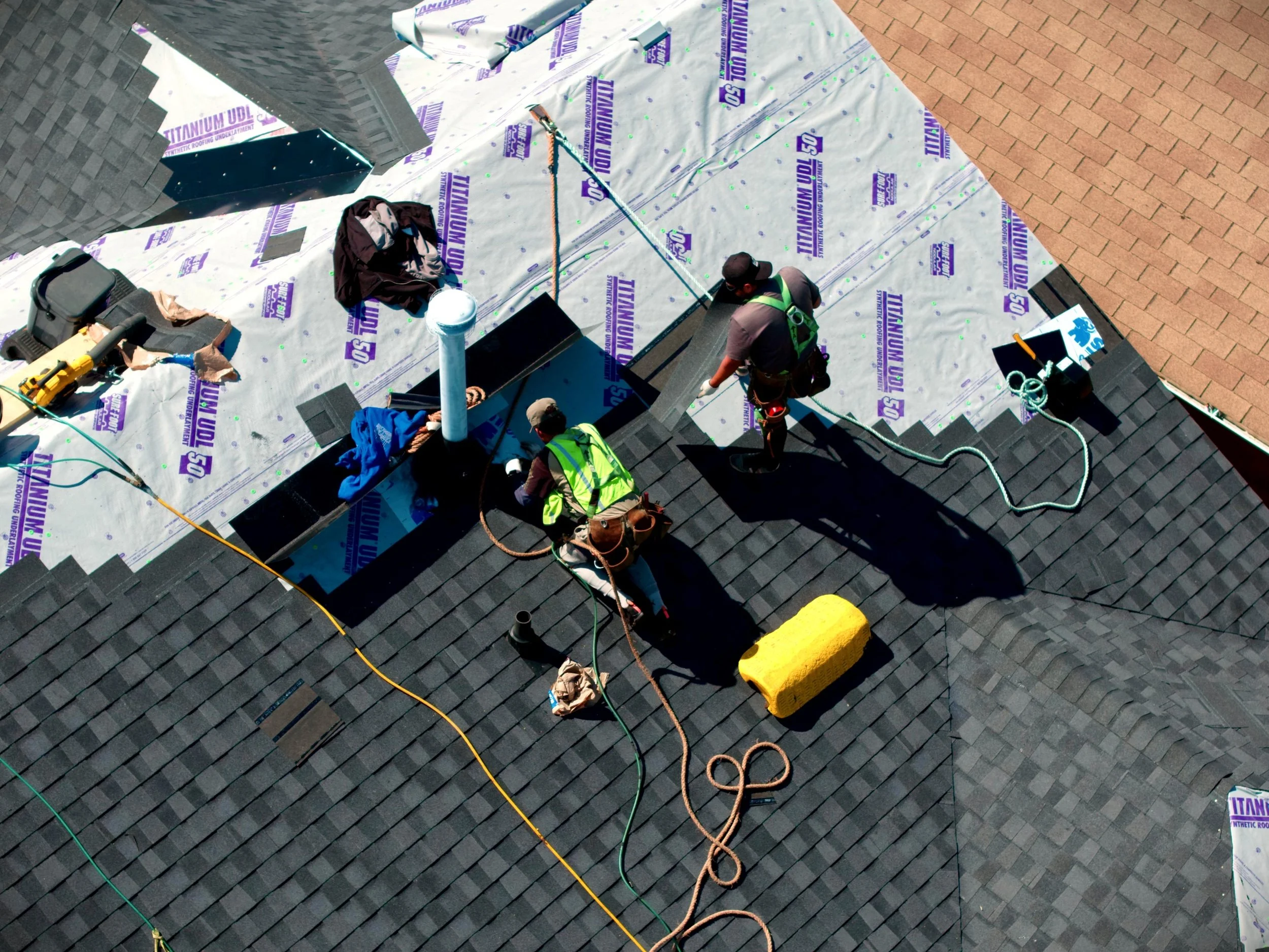 Workers installing shingle roofing on a house, using underlayment labeled 'Titanium UDL 50'. They are working on the roof with tools, hoses, and safety gear, with part of the roof covered in black shingles and part in brick-paved area.