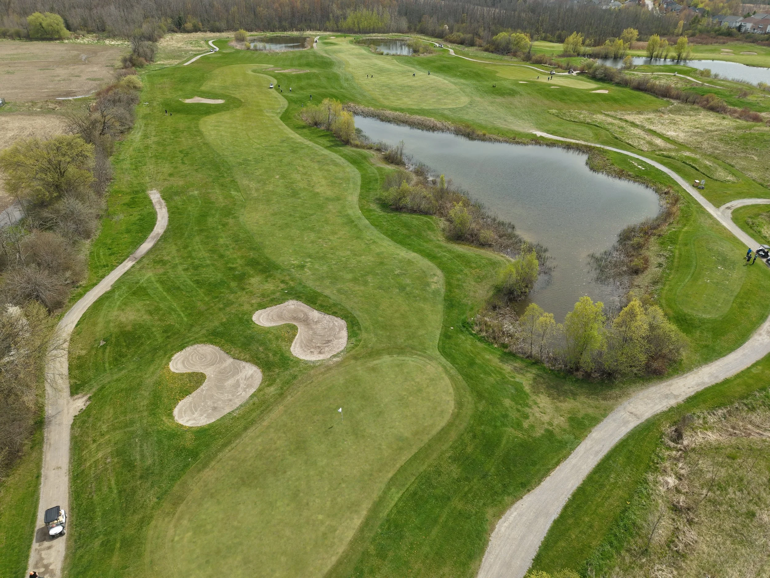 Aerial view of a golf course with several holes, sand traps, water hazards, and winding pathways, surrounded by trees and a few buildings in the background.