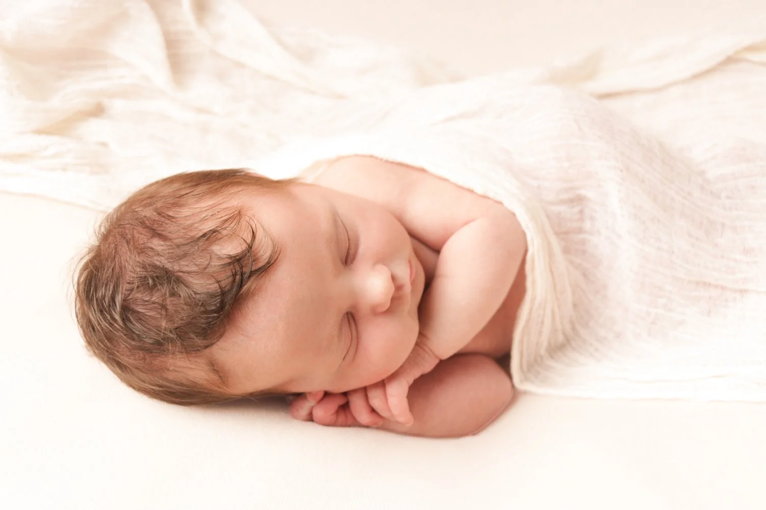 Peacefully sleeping newborn baby with dark hair wrapped in a soft cream blanket, photographed in a natural neutral newborn photography studio in Staffordshire.