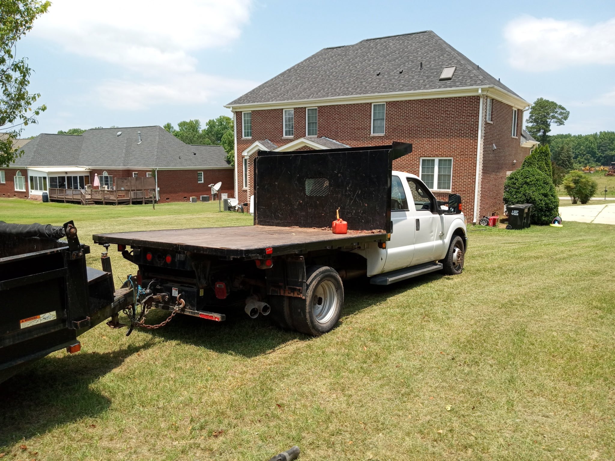 A residential backyard with a large two-story brick house, a wooden deck, and a green lawn. There are two trucks in the foreground, one with a black flatbed and the other with a black cover. The background shows trees and neighboring houses.