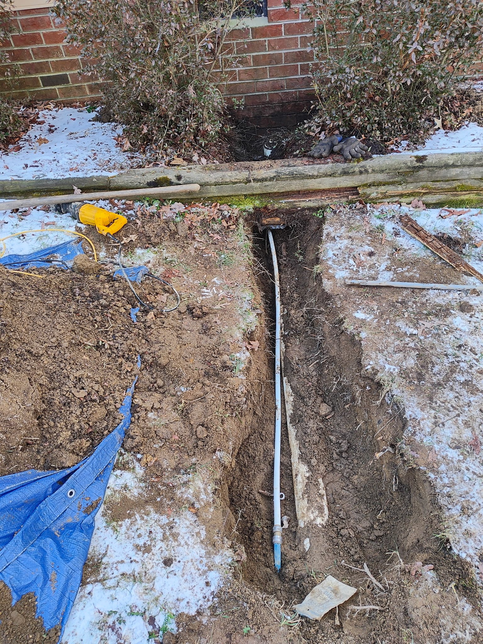Underground pipe installation in a yard with dirt, snow, and construction tools including a yellow electric drill, a blue tarp, gloves, and pipes.