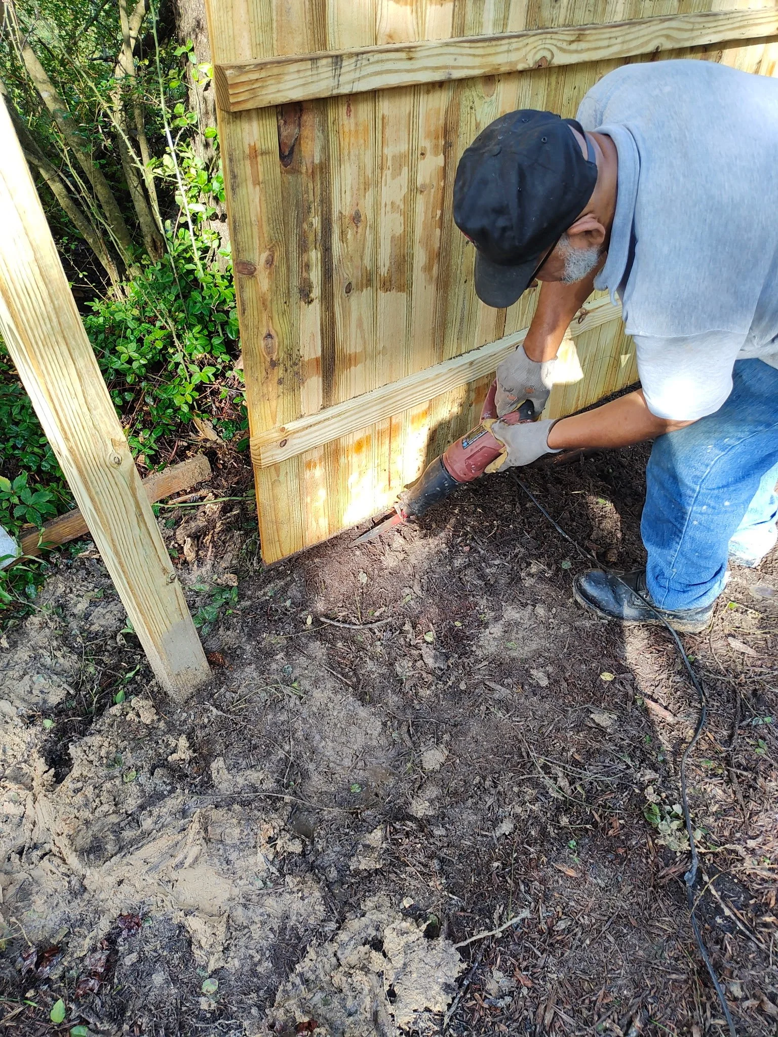 A person working on a wooden fence outdoors, using a power screwdriver to attach a horizontal wooden panel to the vertical posts. The person is wearing gloves, a gray shirt, a black cap, and jeans, and appears to be focused on the task.