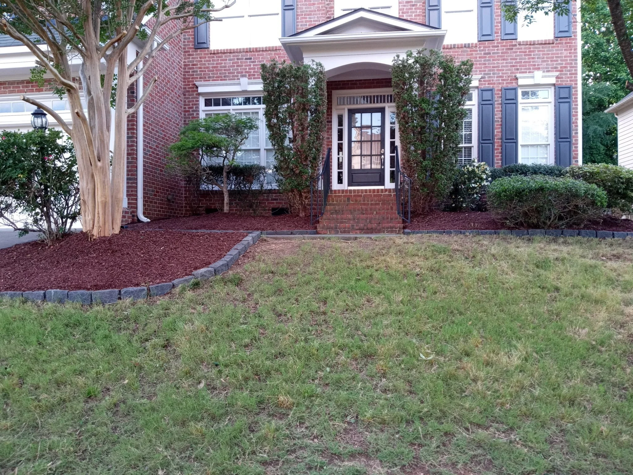 Front view of a brick house with steps leading to the front door, surrounded by landscaped yard with trees and bushes.