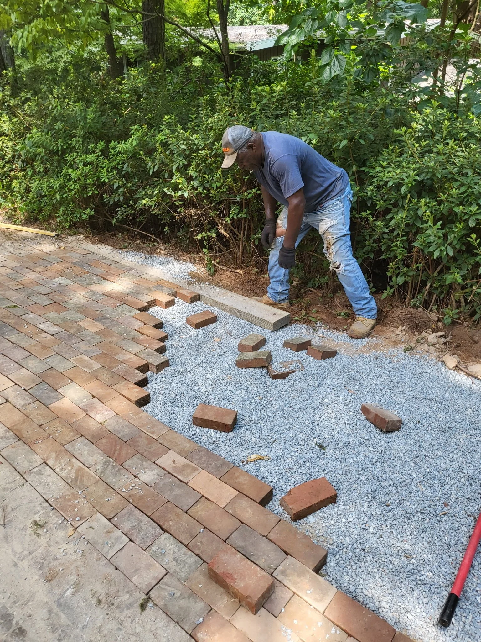 A person laying brick pavers on a gravel base in a backyard garden, with green bushes, trees, and a shed in the background.