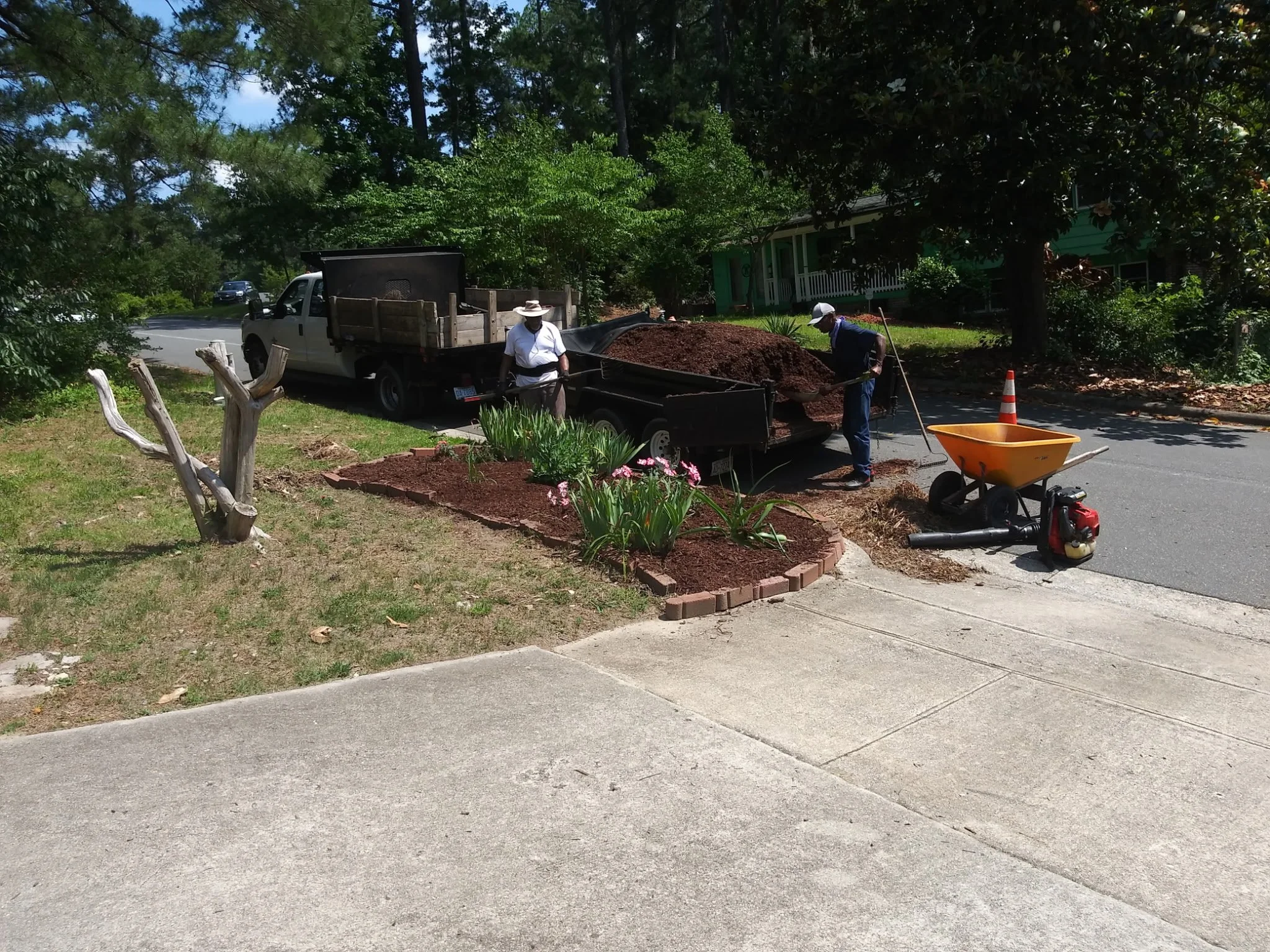 Workers spreading mulch in a landscaped area near a sidewalk, with a pickup truck and various gardening tools present.