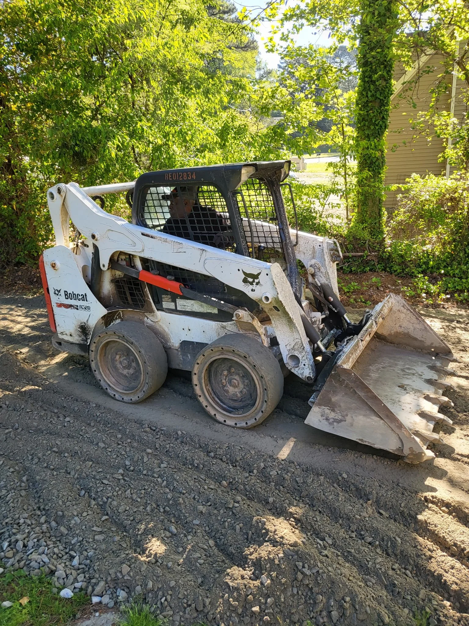 A small white Bobcat skid-steer loader with a black cage around the operator's seat is parked on a gravel surface beside a bush and a tree with green leaves. The loader has a shovel attached at the front, partially resting on the ground.