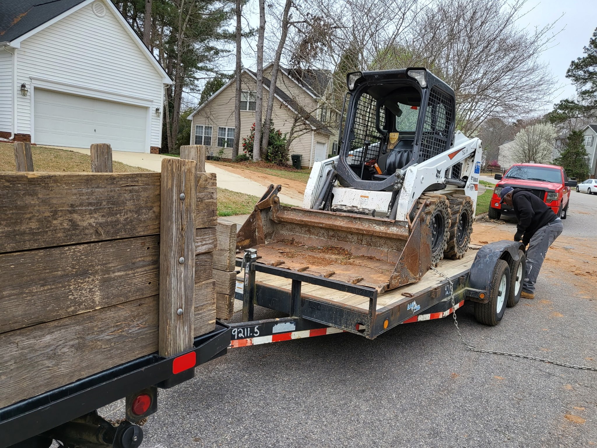 A flatbed trailer loaded with wooden boards and a small skid-steer loader parked on a residential street with houses and trees in the background.
