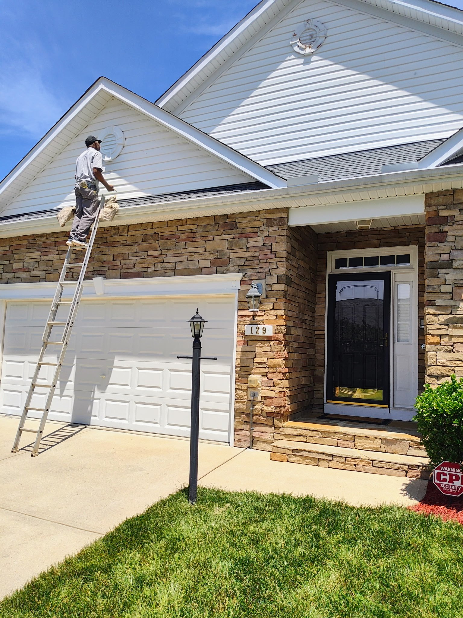 A man standing on a ladder cleaning gutters of a white house with brick accents, a black door, and a white garage door with a dark roof. The house has house numbers 129 and a security sign in the front yard, with a lawn and a sidewalk under a clear blue sky.