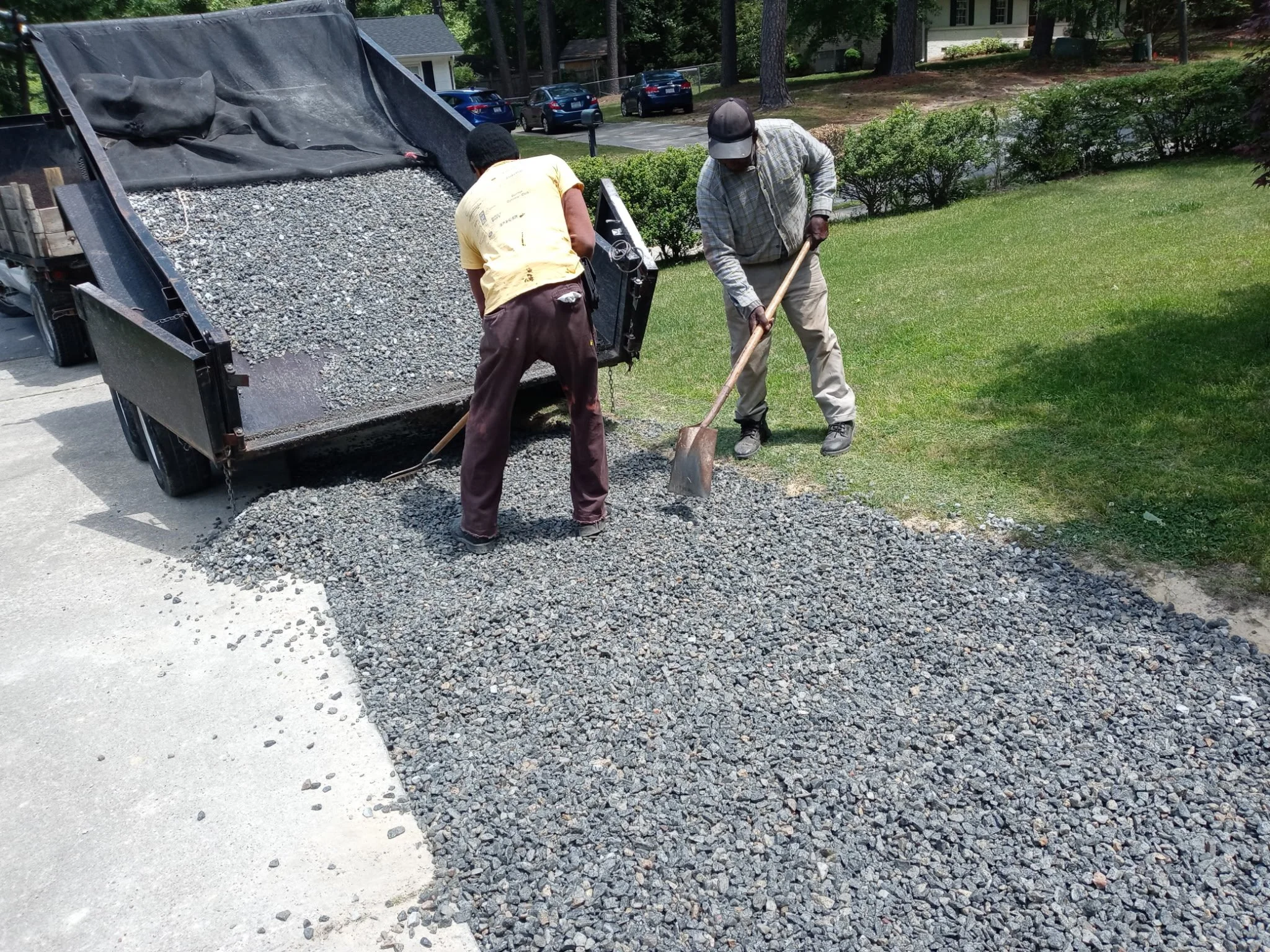 Two men are spreading gravel on a driveway next to a trailer filled with gravel. One man is using a shovel, and the other is spreading gravel with a tool.