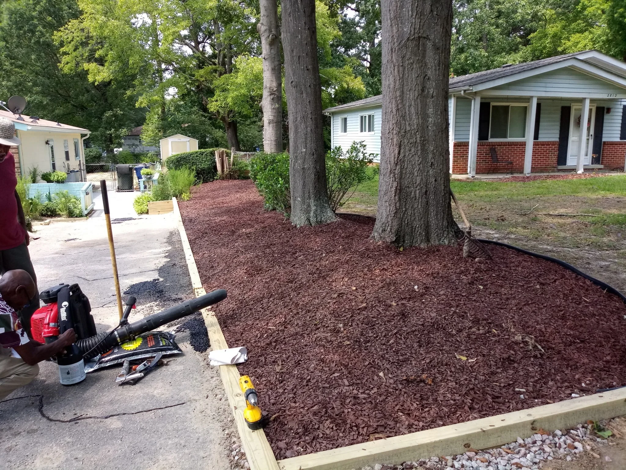 Individuals working on landscaping or garden bed installation, with fresh mulch, large trees, a house, and garden tools visible.