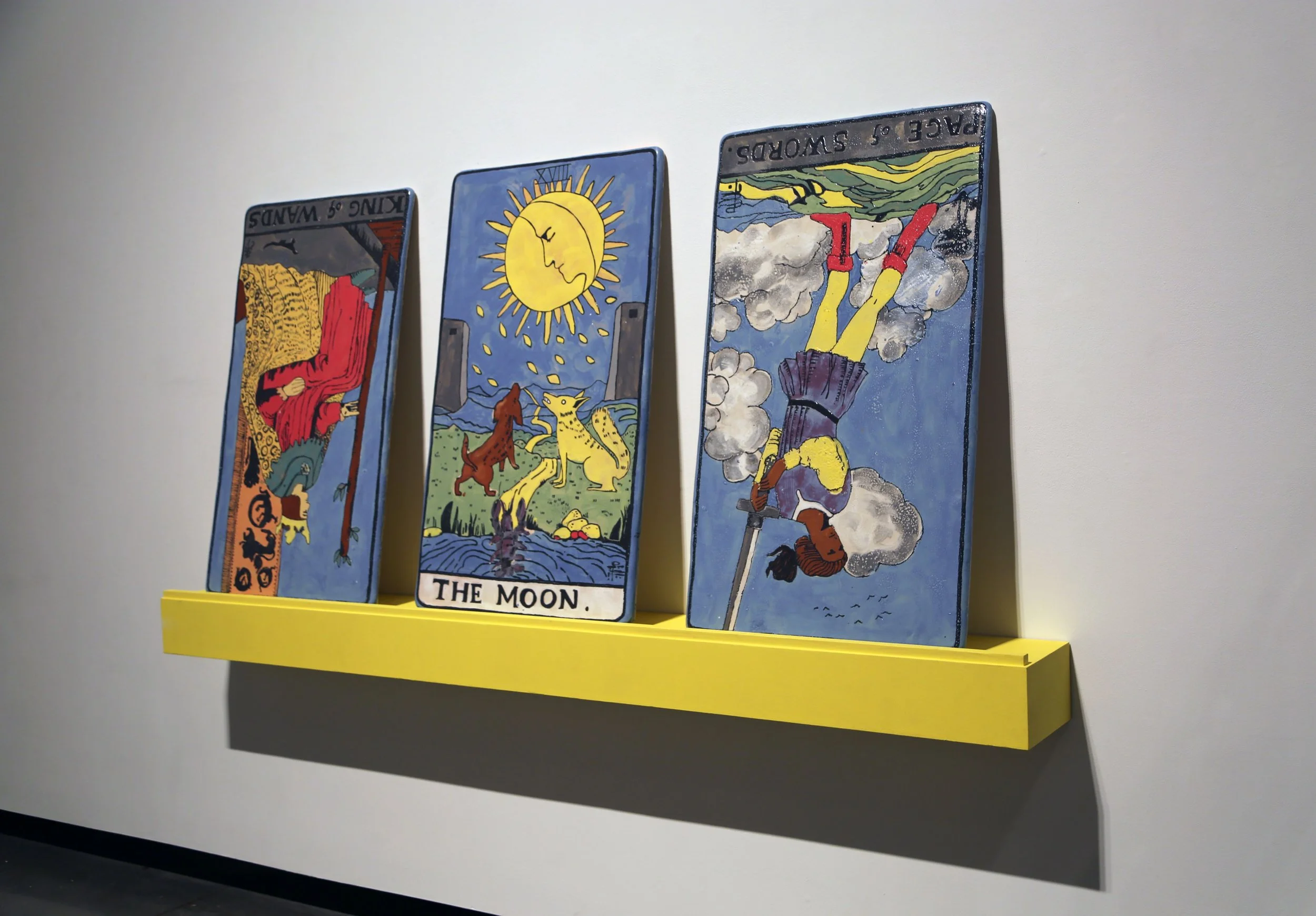 Three tarot cards displayed on a bright yellow shelf against a plain wall. The cards include The Moon, the Two of Swords, and the King of Wands, each with colorful, illustrative artwork.