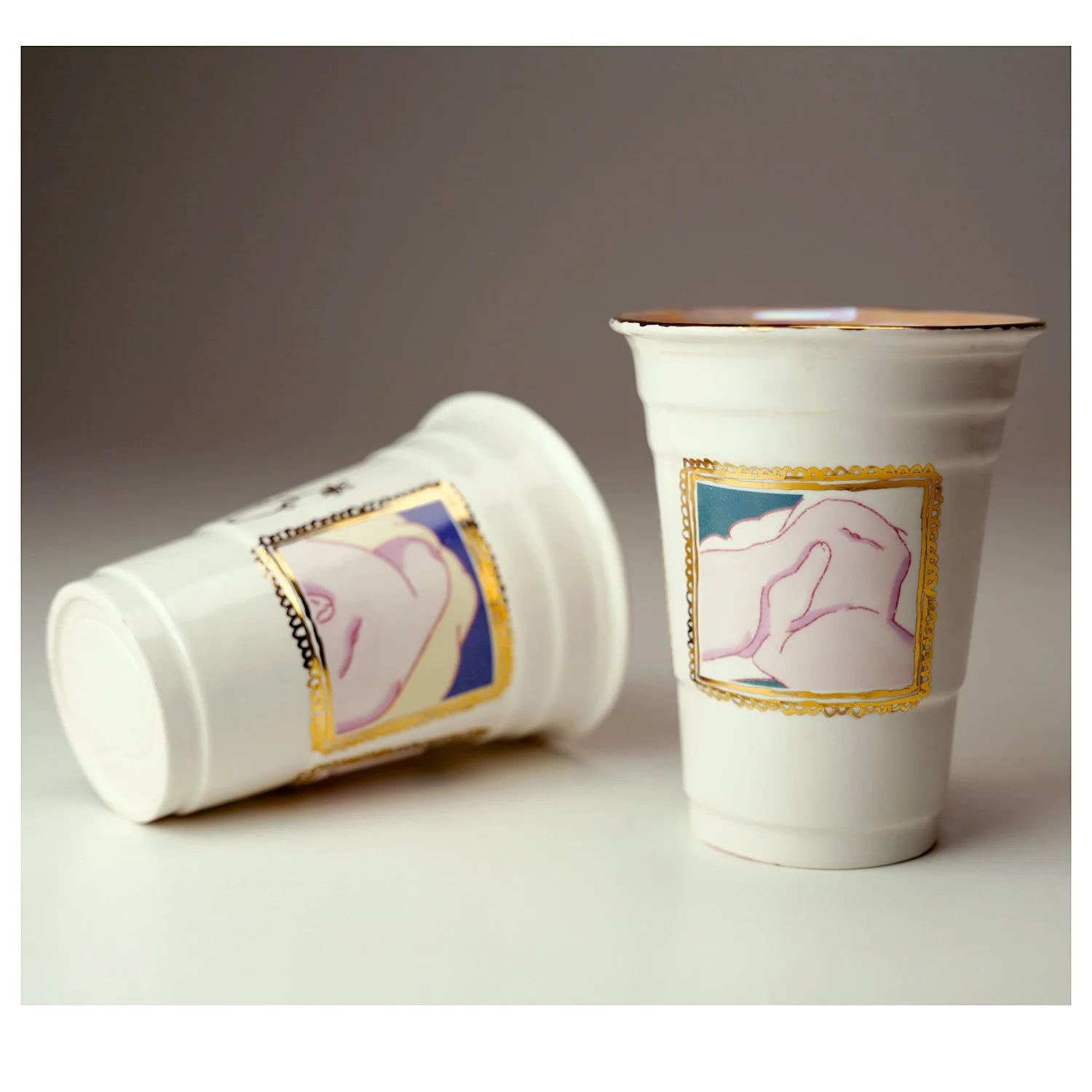 Two disposable cups with a colorful graphic label placed on a plain surface. One cup is upright, and the other is lying on its side, both featuring a design with abstract pink and blue artwork and a gold border.