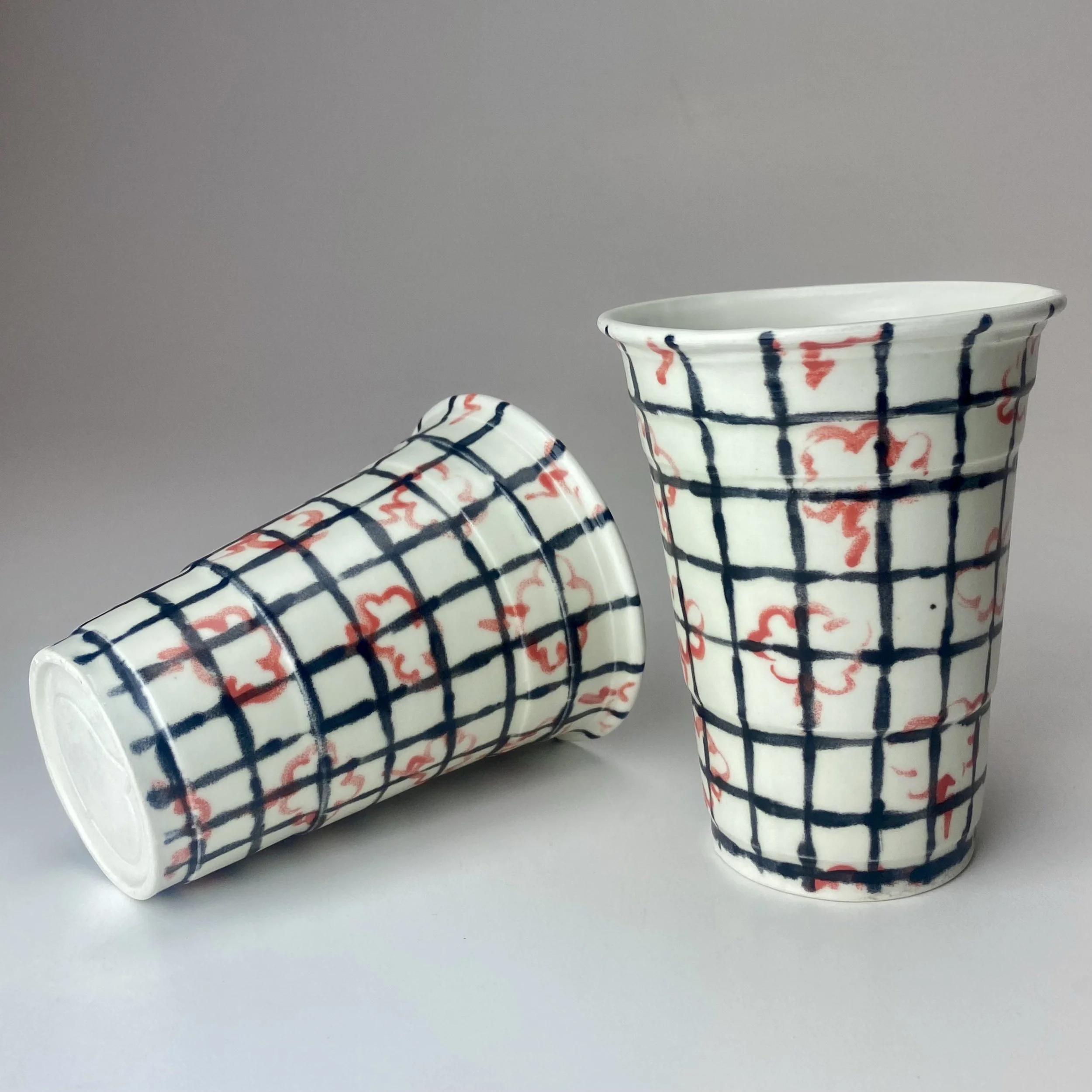 Close-up of three ceramic cups with colorful red and blue grid and floral patterns, placed on a light gray surface.
