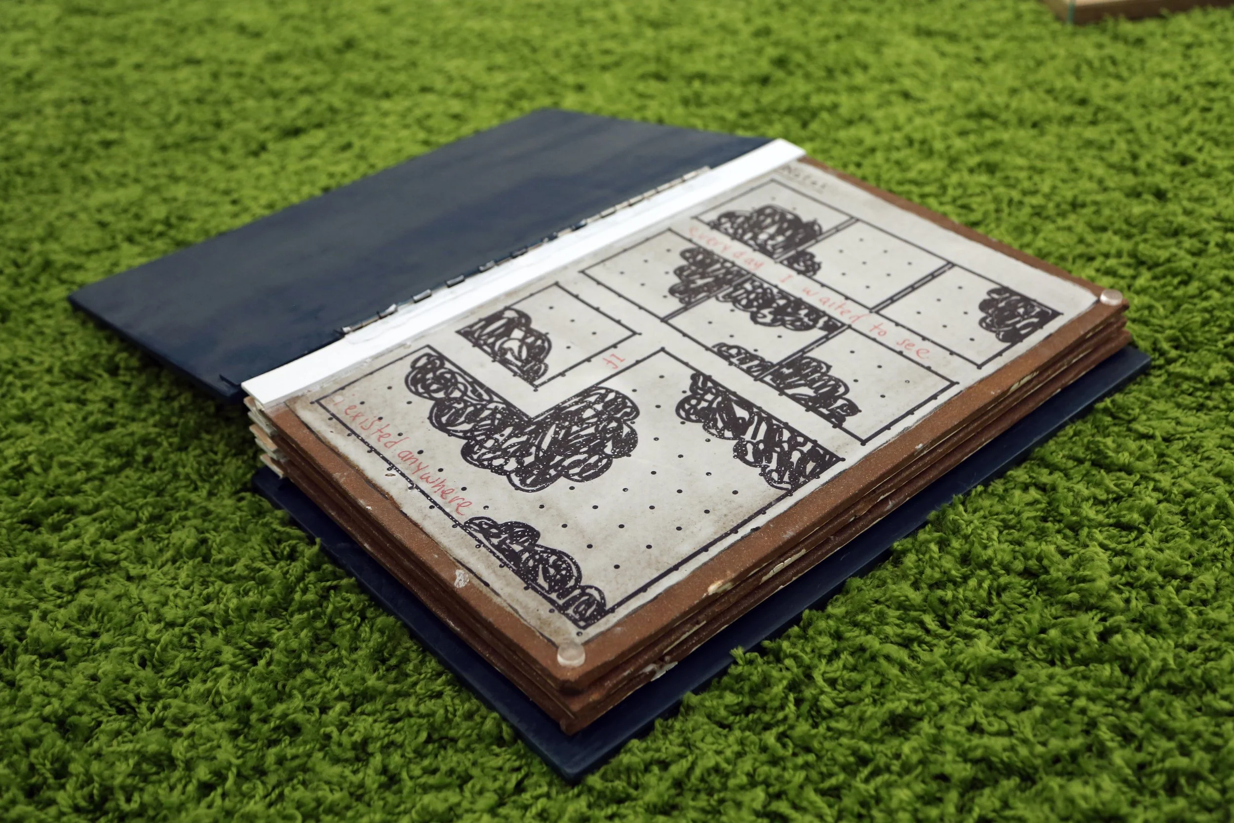 A scrapbook open on a grassy surface, showing a floor plan drawing with black scribbles and red handwritten notes.
