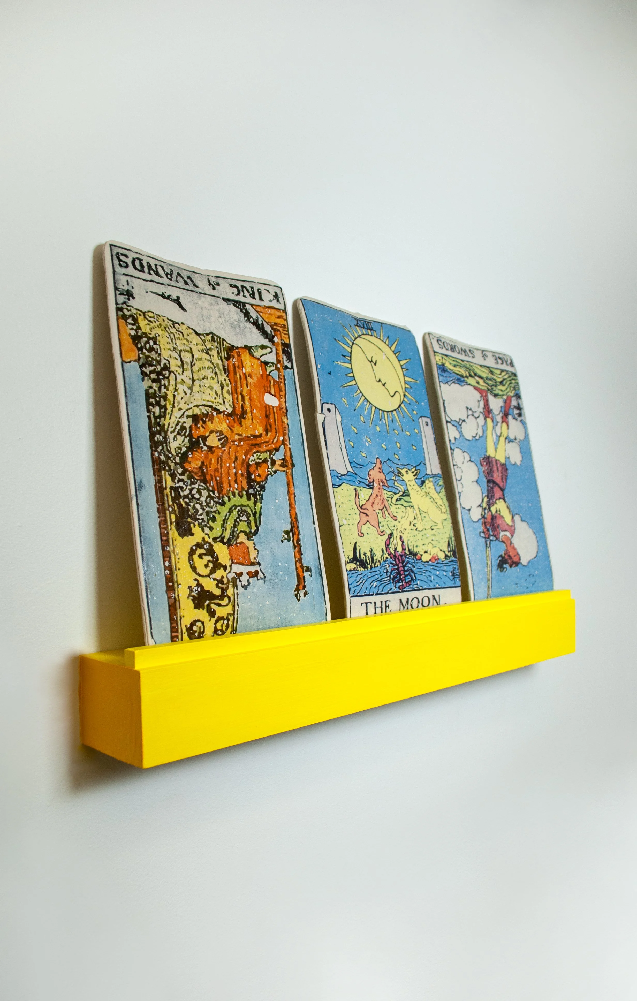 Three tarot cards — The World, The Moon, and The Sun — displayed on a yellow wall shelf, against a white background.