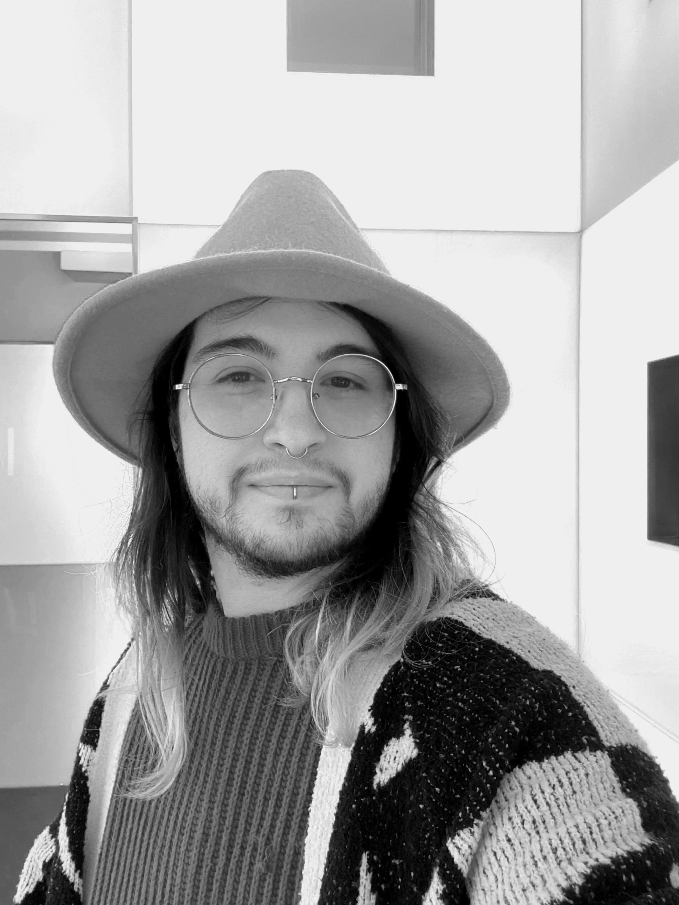 A person with long hair, glasses, and a septum piercing wears a wide-brim hat and a patterned sweater, taking a selfie in an indoor setting.